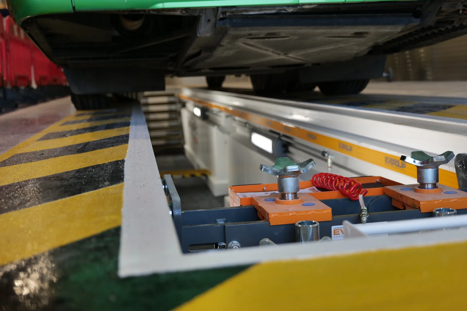 Underneath view of a green vehicle on a lift with orange support fixtures, yellow and black striped safety markings, and metallic tools and components.