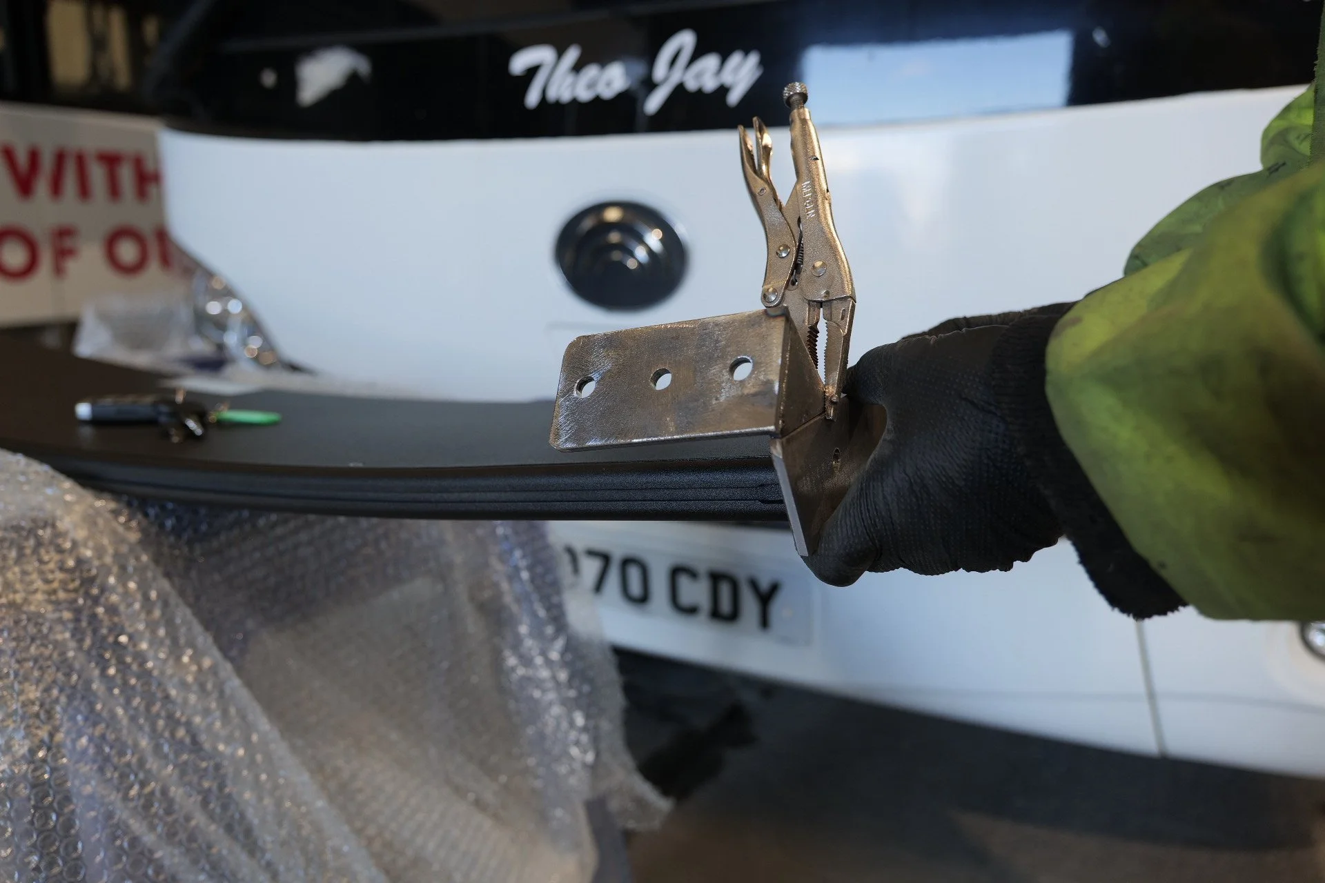 A person working on a vehicle, seen holding a metal hinge or bracket part, with a white vehicle and license plate in the background.