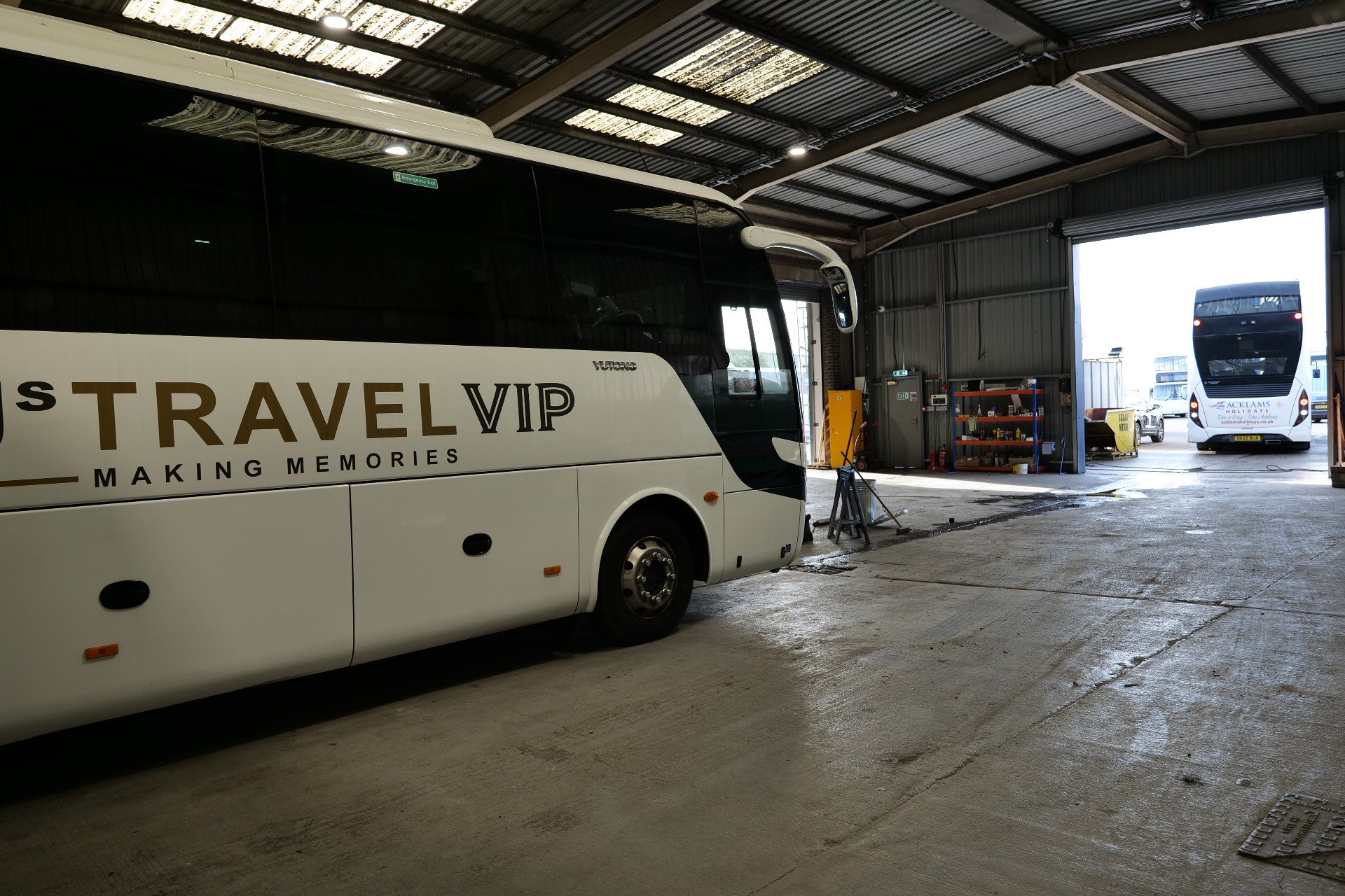 Inside a large garage or bus depot with two buses visible, one labeled 'Travel VIP' in the foreground and another in the background, both parked indoors near open garage doors.