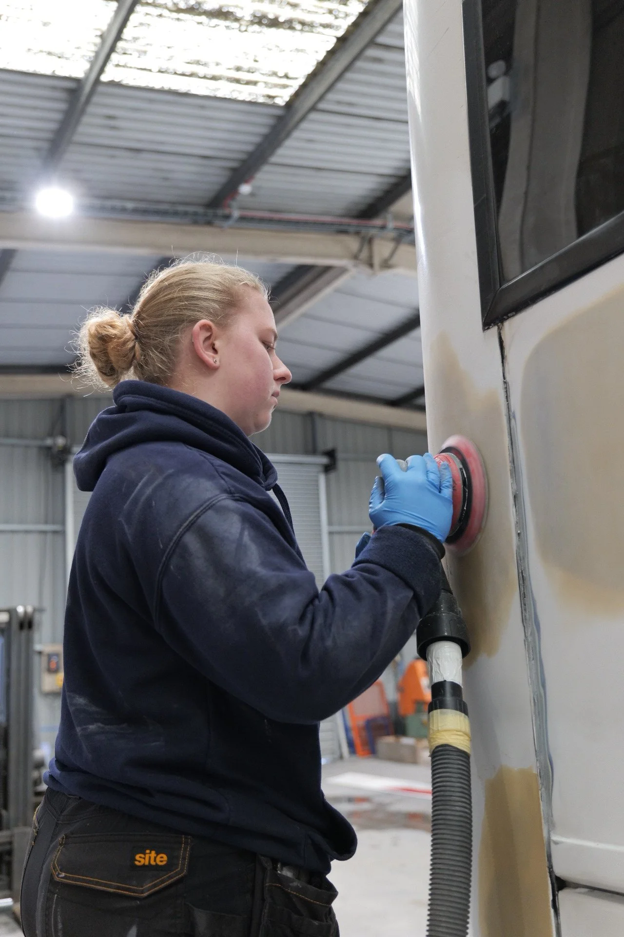 A woman with blonde hair tied in a bun, wearing a navy hoodie, using a power buffer on the side of a cream-colored vehicle inside a workshop.