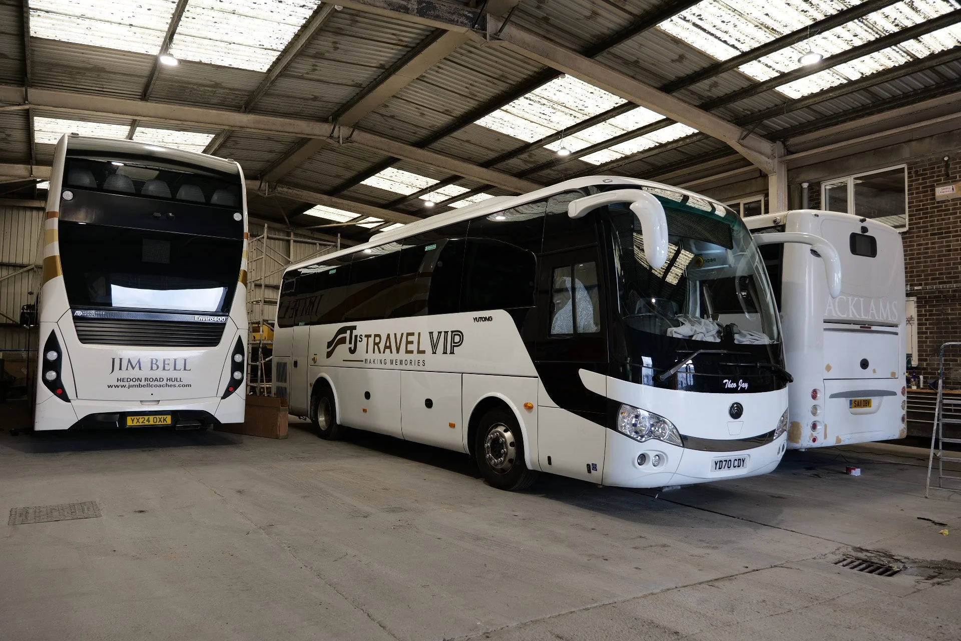 Three buses parked inside a warehouse or garage, with a metal roof and brick wall in the background. The buses are white with black accents and branding.