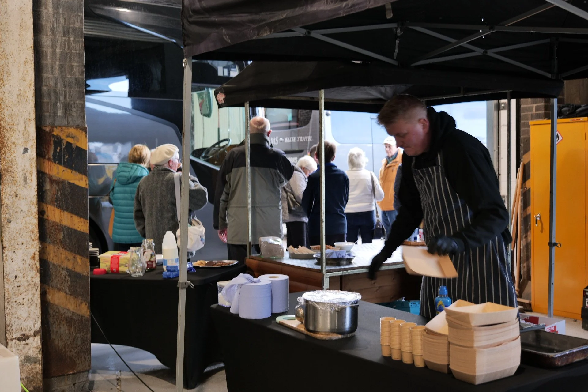 A street food stall with a man preparing food, surrounded by a group of people waiting or buying, parked buses in the background.