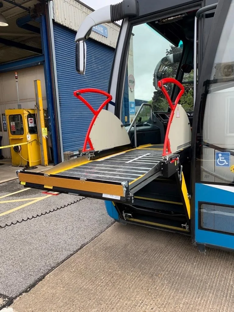 Accessible bus with wheelchair lift extended at the entrance, parked outside a facility with a blue rolling door and a yellow payment kiosk nearby.