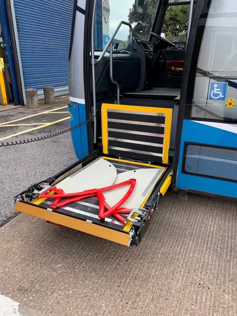 The front door of a blue transit bus is open, revealing the bus interior and a wheelchair accessible ramp extended to the ground. On the ramp, there are bright red safety barriers and a white platform, with the bus parked in a lot near a blue rolling shutter.