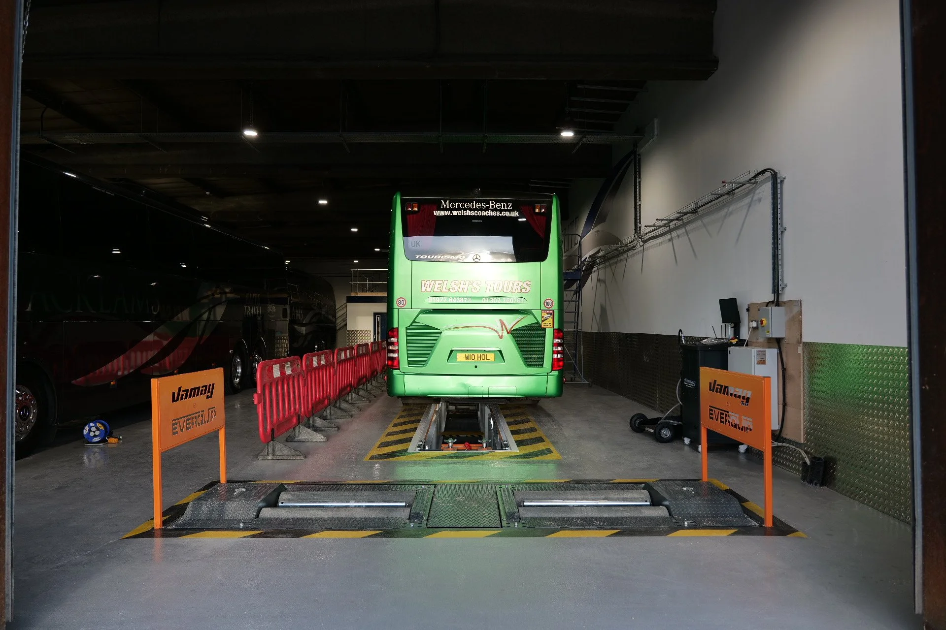 Interior of a vehicle inspection or repair garage with a green double-decker bus on a testing or lifting platform, surrounded by orange safety barriers and equipment.