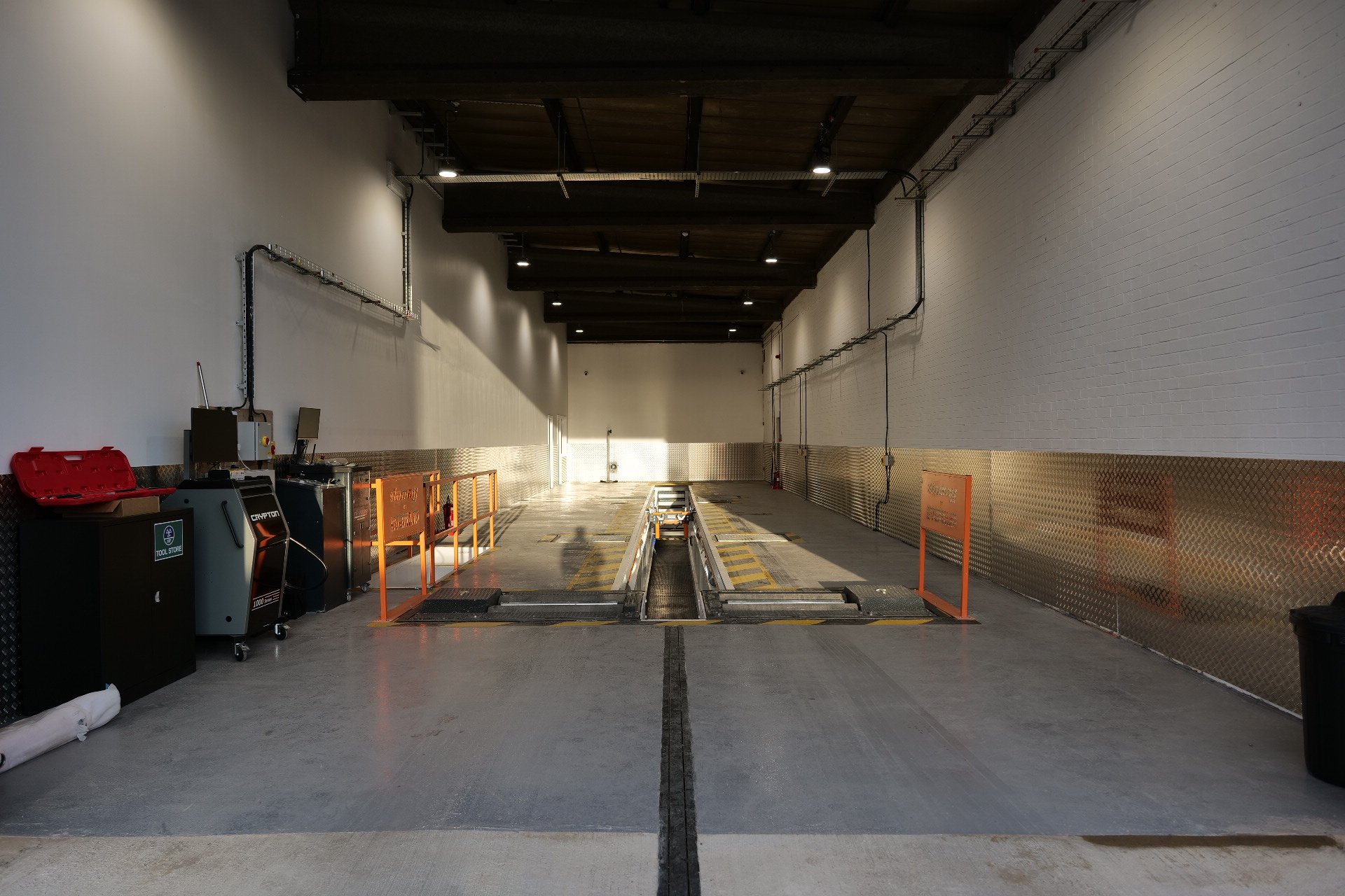 Empty vehicle inspection pit in a garage or workshop with machinery and tools on the left side and a painted wall on the right.