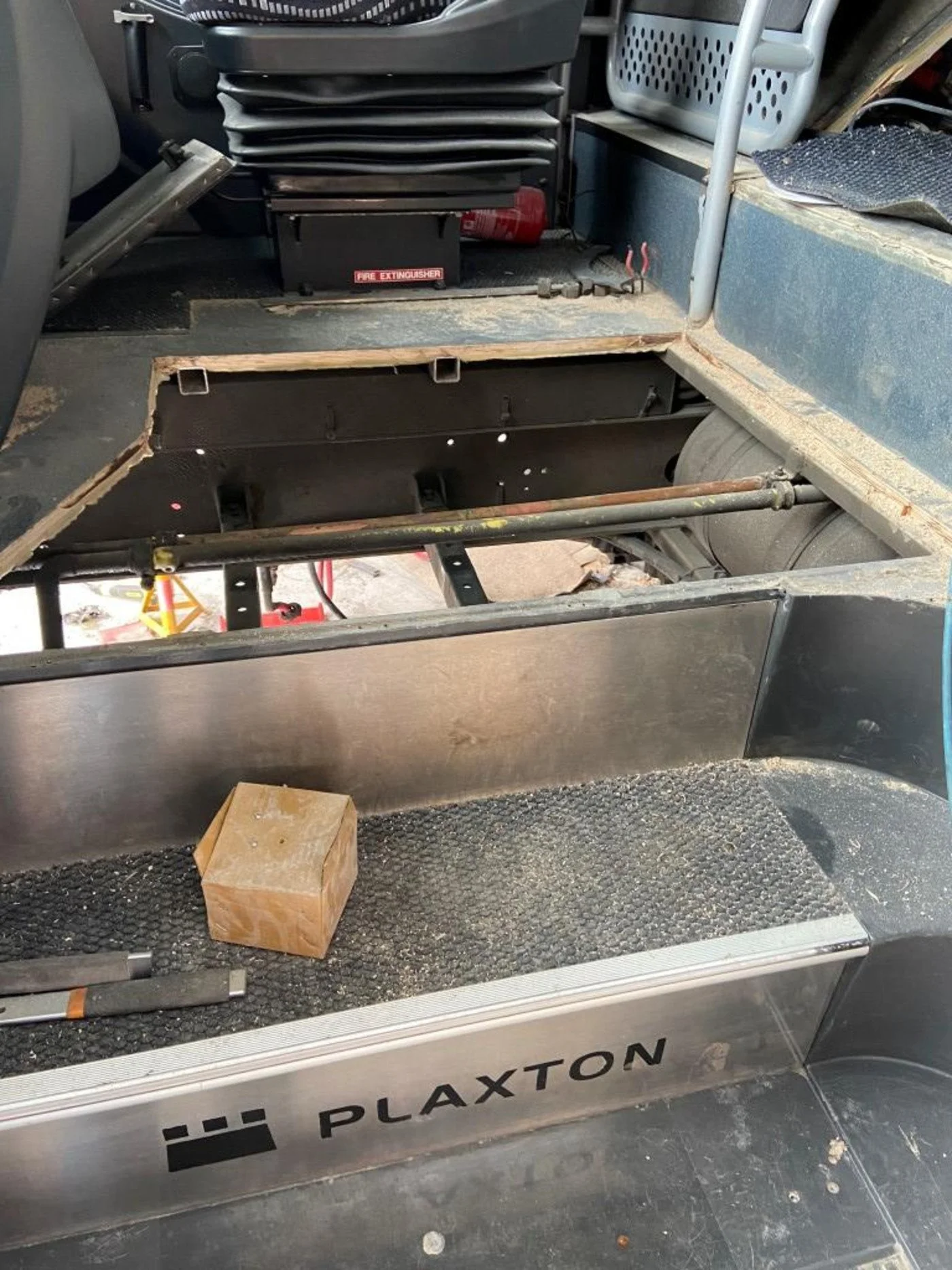 Interior of a vehicle undergoing repair with missing flooring panel, exposing metal framework and steering components. Tools and a wooden block are on the floor. The vehicle has a Plaxton logo.