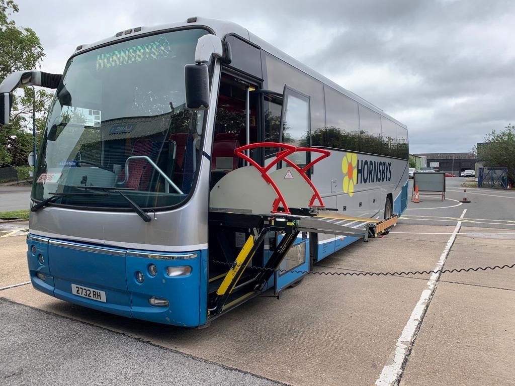 A large gray and blue bus labeled 'HORSENS' parked outside. The bus has one of its side panels removed, revealing the interior and machinery, with orange safety cones in the background.