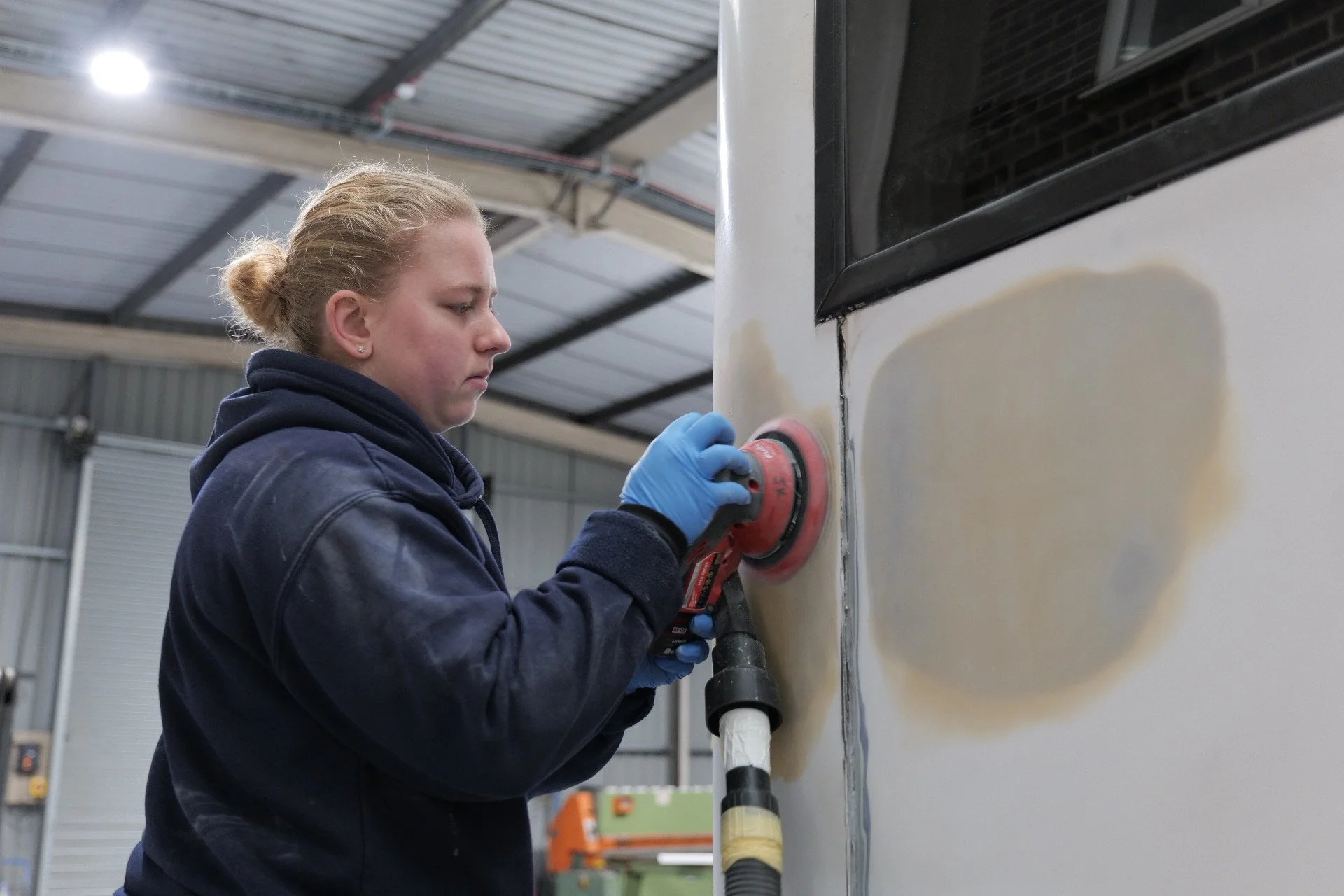A young woman wearing a navy hoodie and blue gloves polishing the side of a boat with a power buffer inside a workshop.