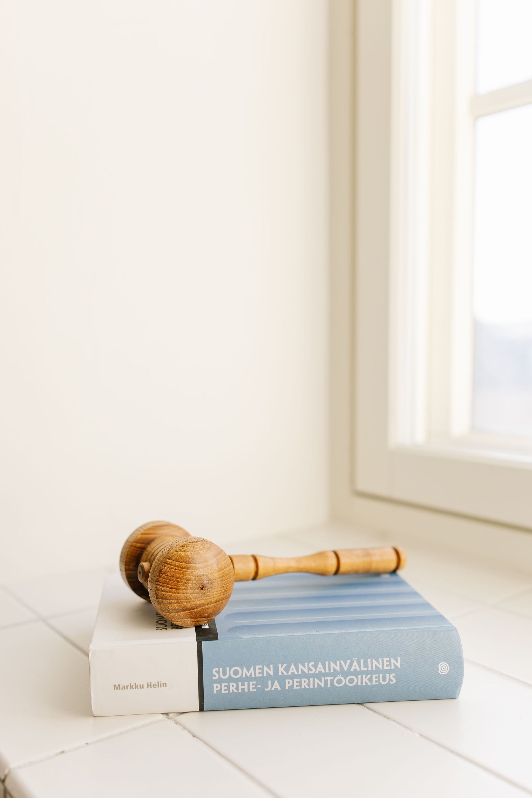 A wooden gavel resting on top of a book titled 'Suomen Kansainvälinen Perhe- ja Perintöoikeus', placed on a white surface near a window.