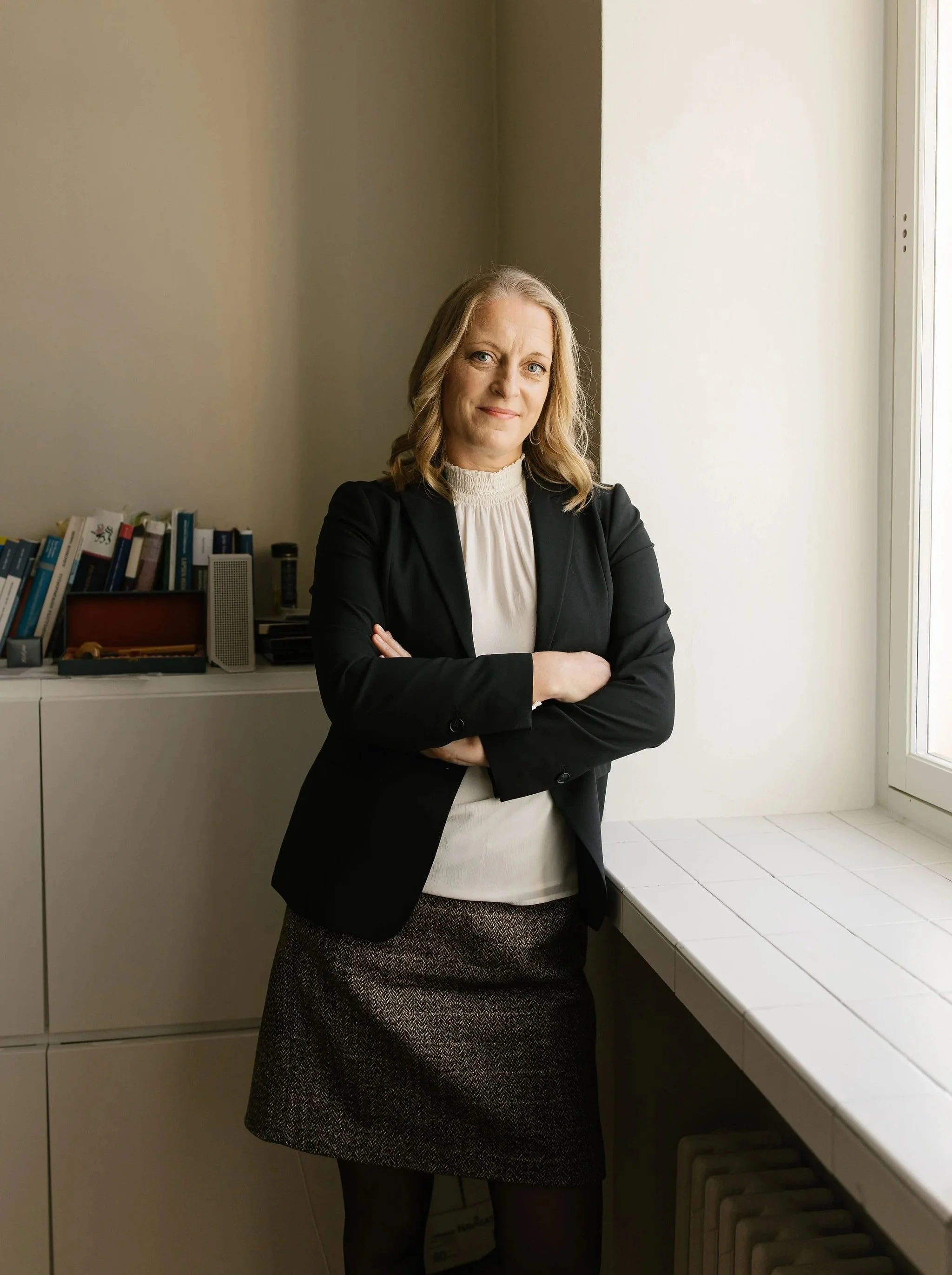 A woman (Lotta Kroneld) standing by a window, wearing a black blazer, white blouse, and skirt, with her arms crossed, in an office setting.