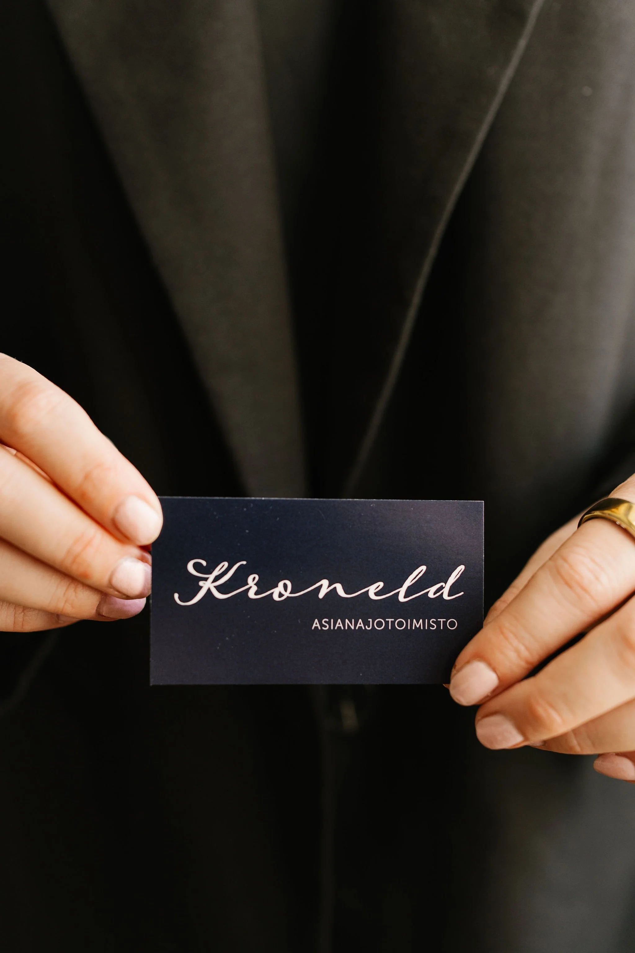Person holding a business card with the name 'Kroneld' and the handle 'ASIANAJOTOIMISTO' in front of a dark blazer.