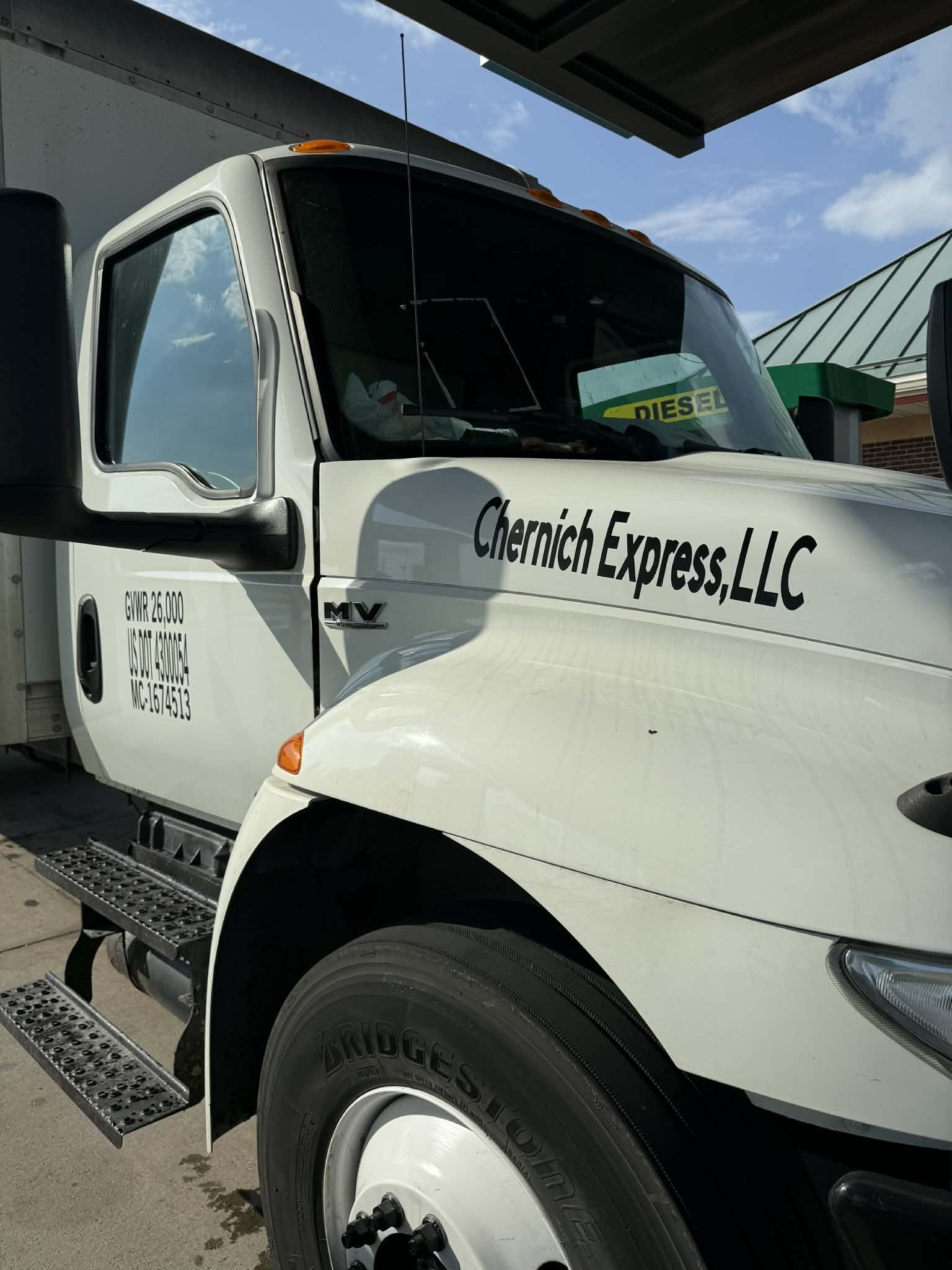 Front side of a white delivery truck with black lettering that reads 'Chernich Express LLC'. The truck has a large front tire, black side mirrors, and a window with a green and yellow sign visible inside.