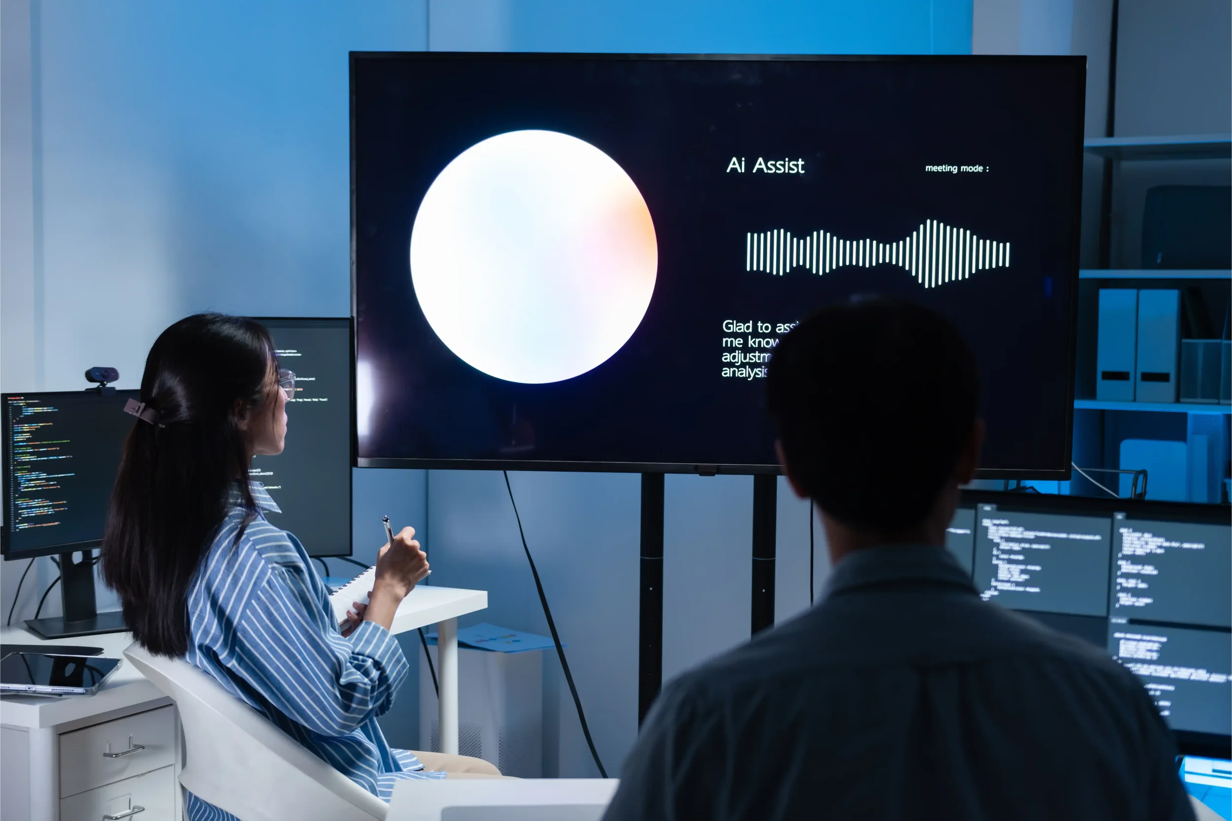 People in a modern office or conference room attending a presentation about AI assist technology, with multiple monitors and a large screen displaying graphics and text.