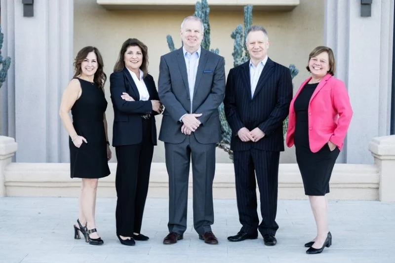 Group of five professionally dressed businesspeople standing outdoors in front of a large cactus and beige building facade.