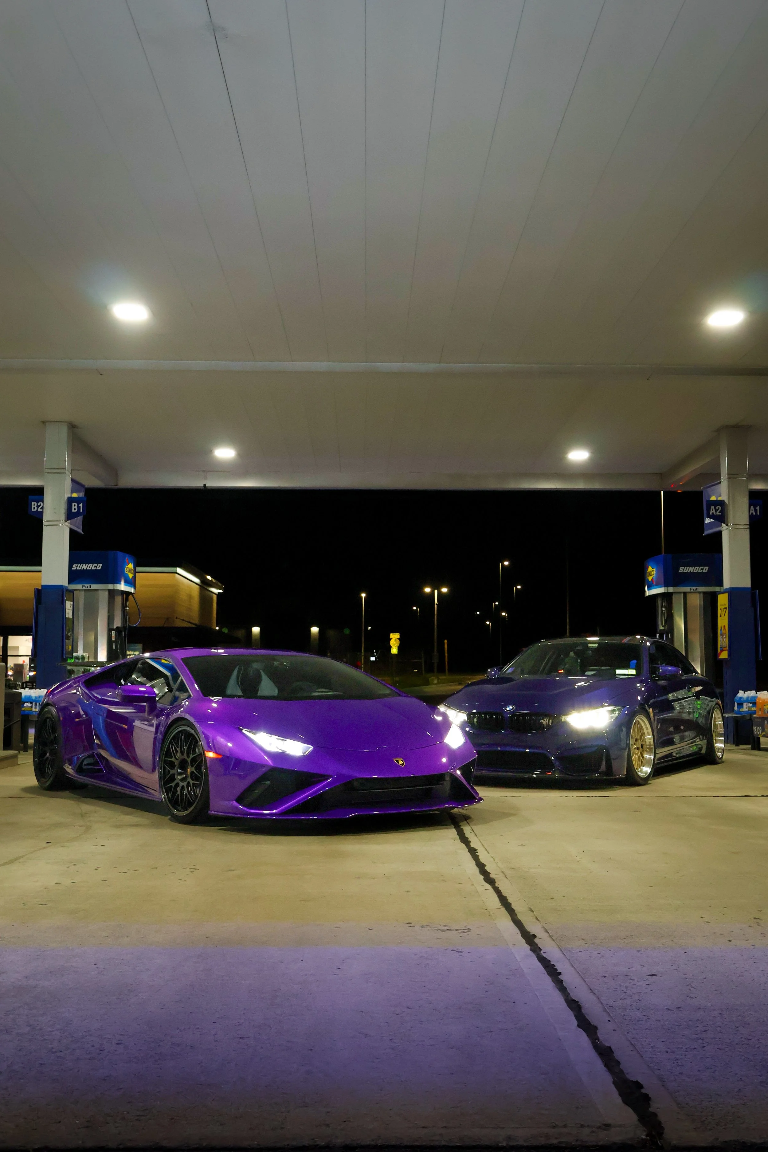 Purple Lamborghini and gray sports car parked at gas station at night under a large canopy with lights; gas pumps visible in background.