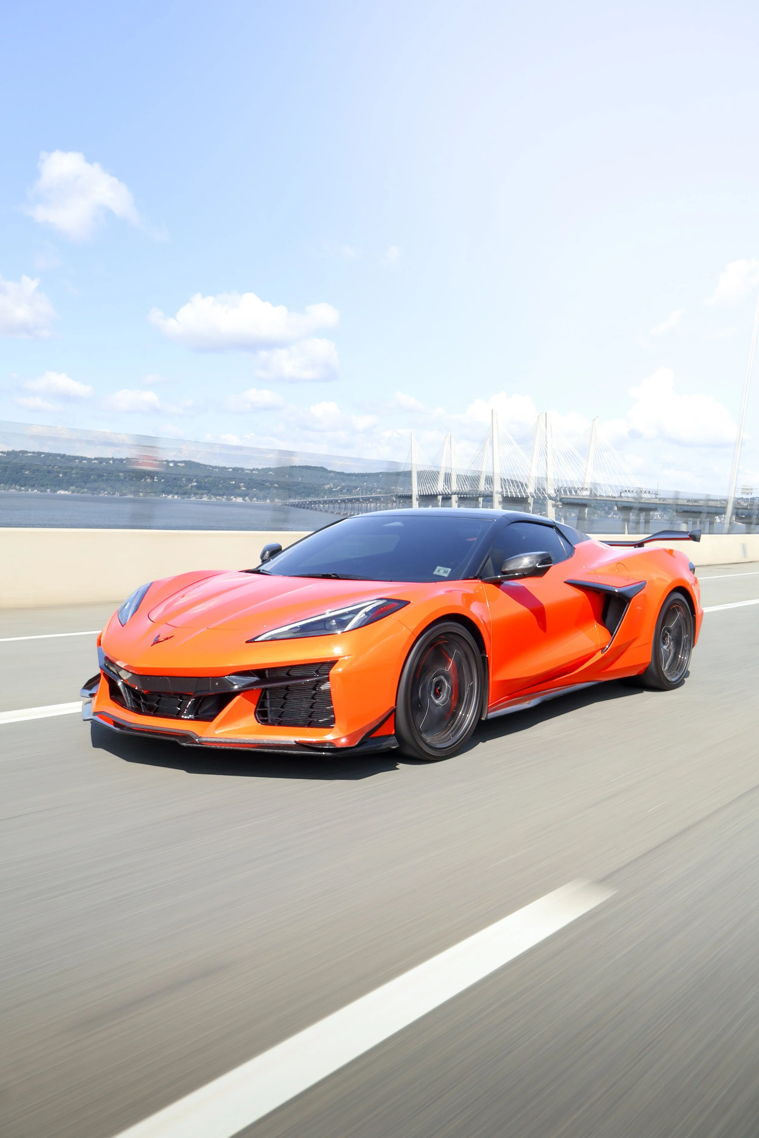 A bright orange sports car with a sleek, aerodynamic design and black accents, driving on a highway under a partly cloudy sky with a bridge visible in the background.