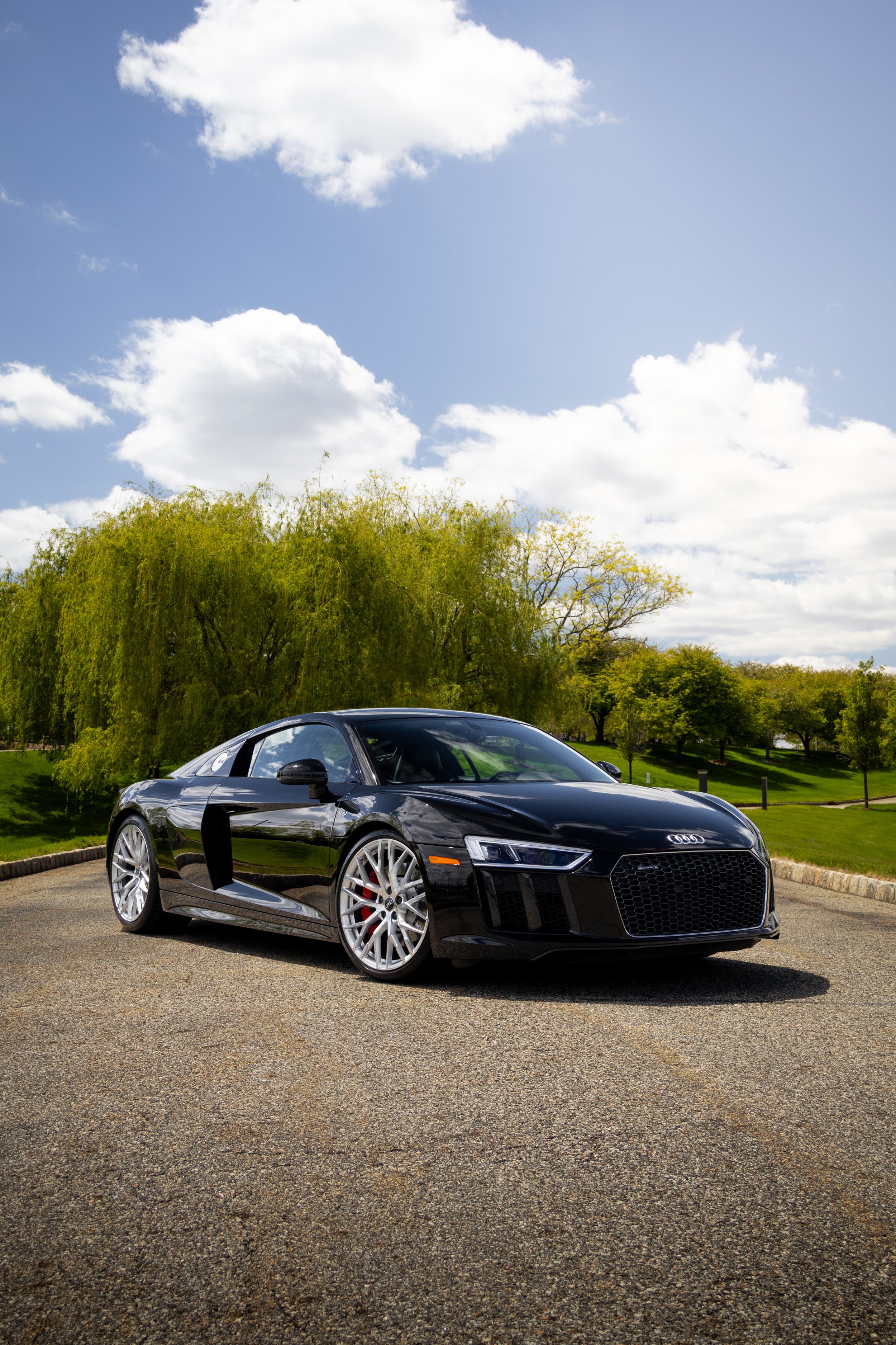 A black Audi sports car parked in an outdoor lot with trees and a partly cloudy sky in the background.