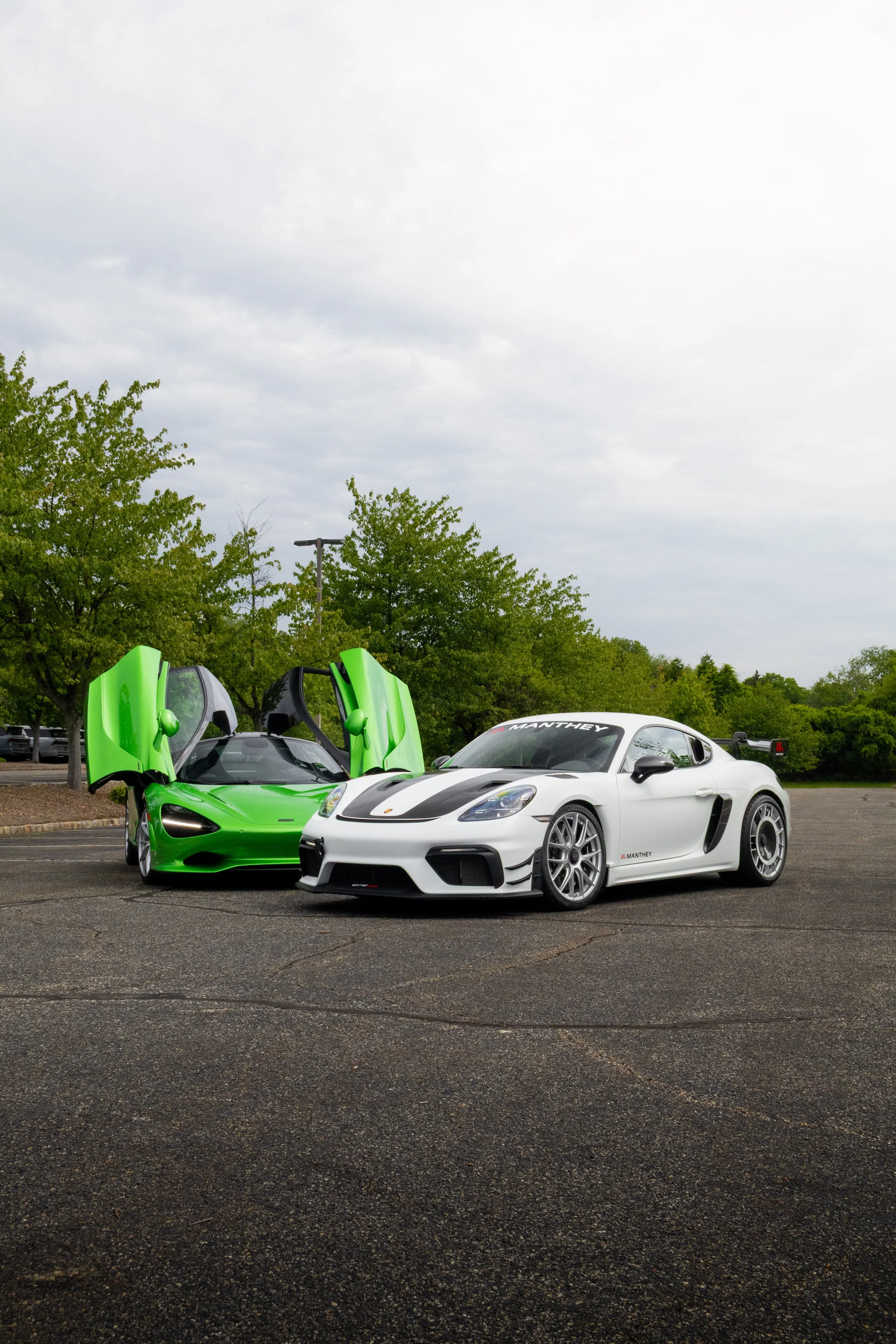 A white sports car and a green sports car with open butterfly doors parked on an asphalt lot, with trees and cloudy sky in the background.