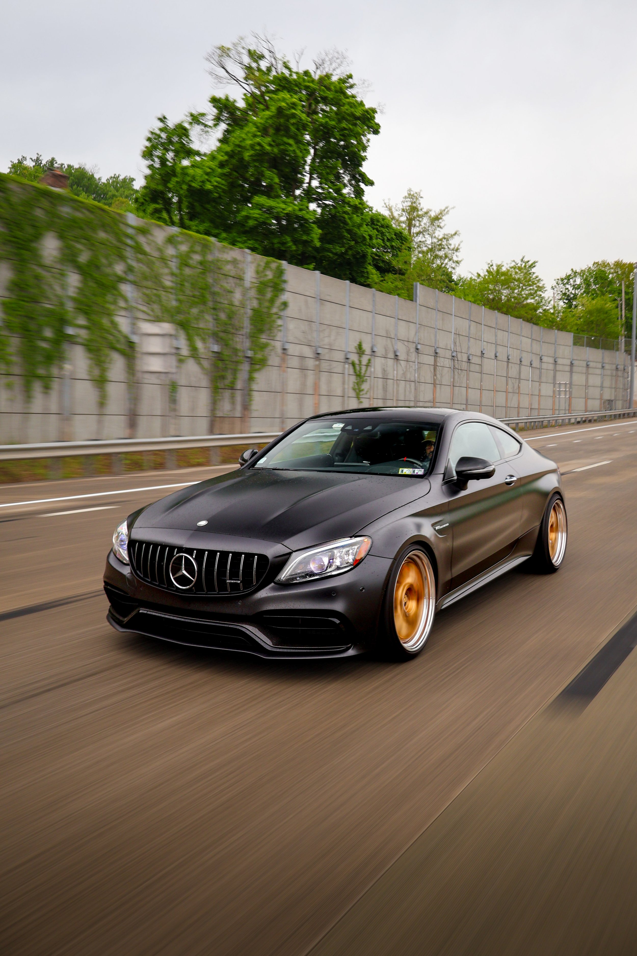 A black Mercedes-Benz coupe with gold rims driving on a highway with a concrete sound barrier and green trees in the background.