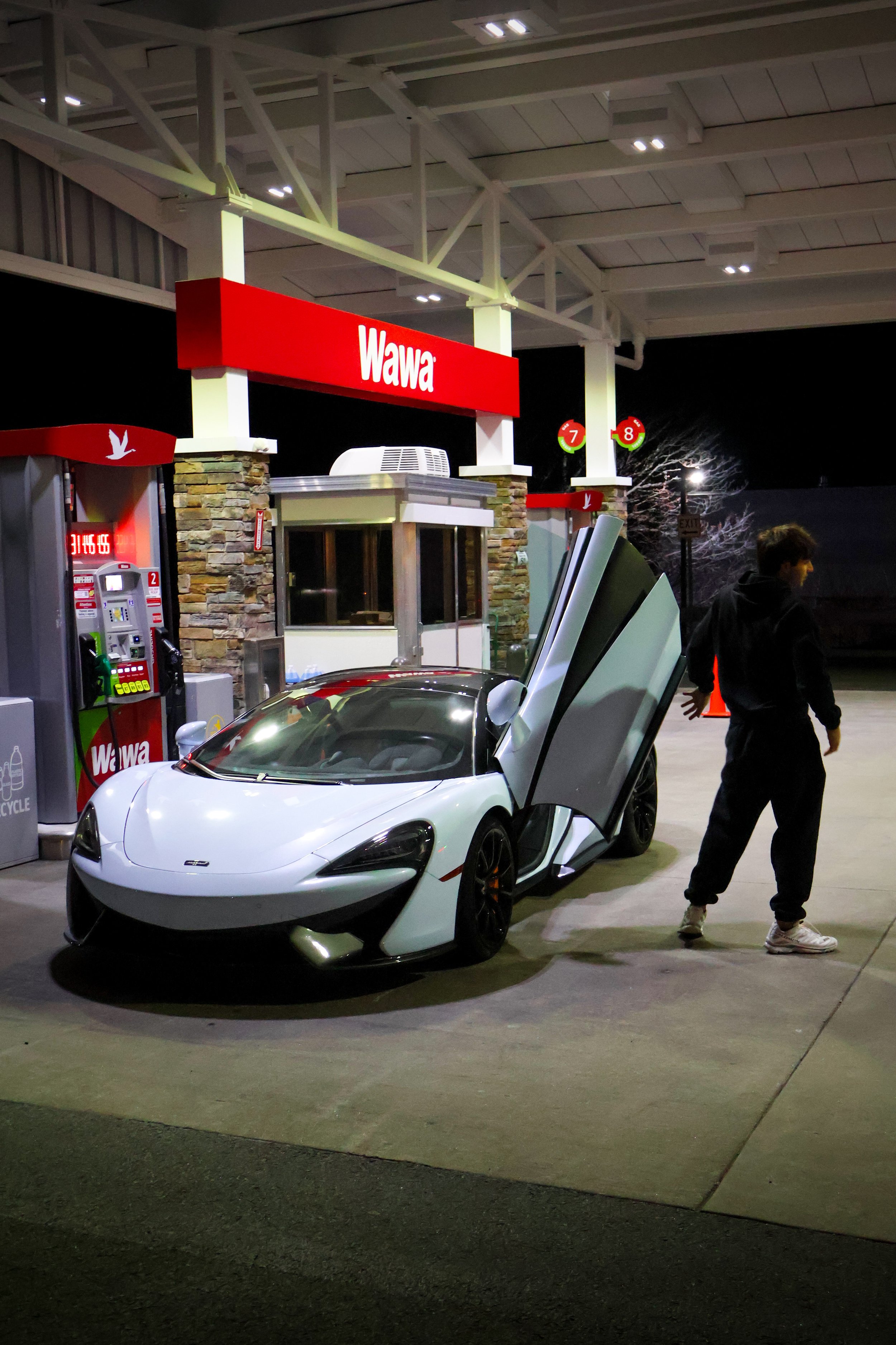 A white sports car with butterfly doors open at a Wawa gas station at night. A person dressed in black with white sneakers is standing next to the car. The gas station has a red sign with white letters that read "Wawa" and a service window with a sto