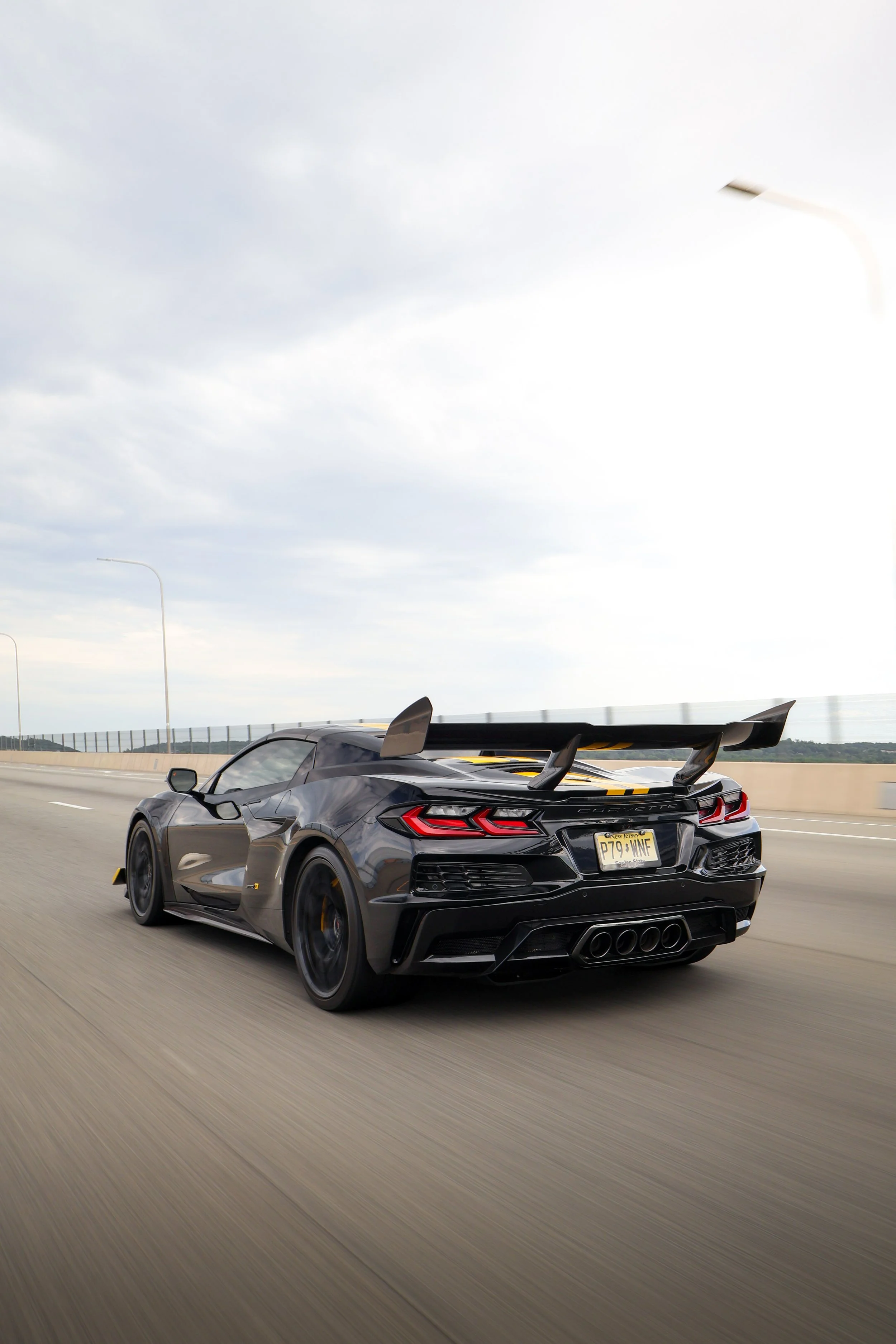 A sleek black sports car with a large rear wing spoiler driving on a highway under a cloudy sky, with a New Jersey license plate that reads P79 WNF.