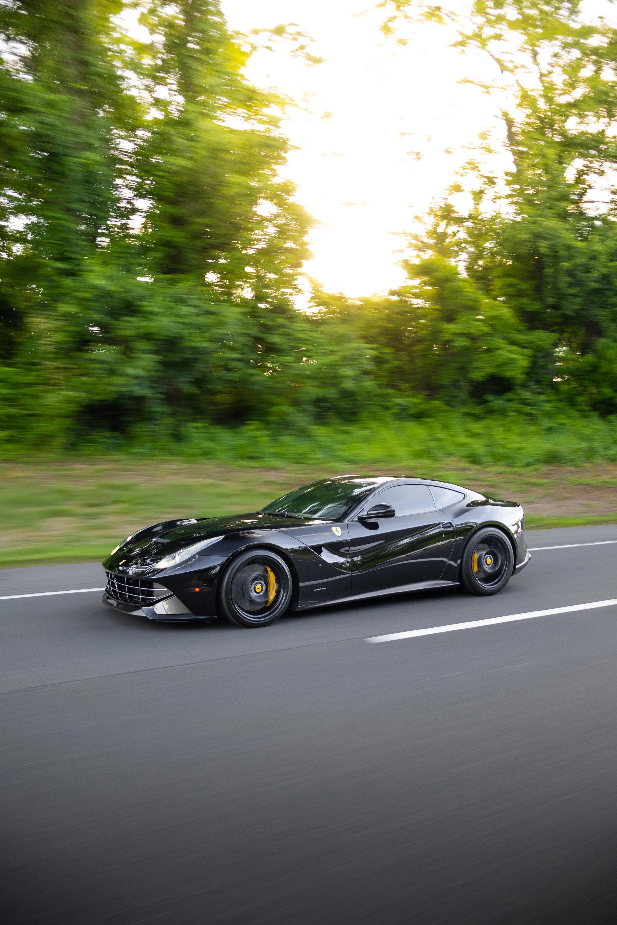A black Ferrari sports car driving on a highway with green trees on the side and sunlight shining through the leaves.