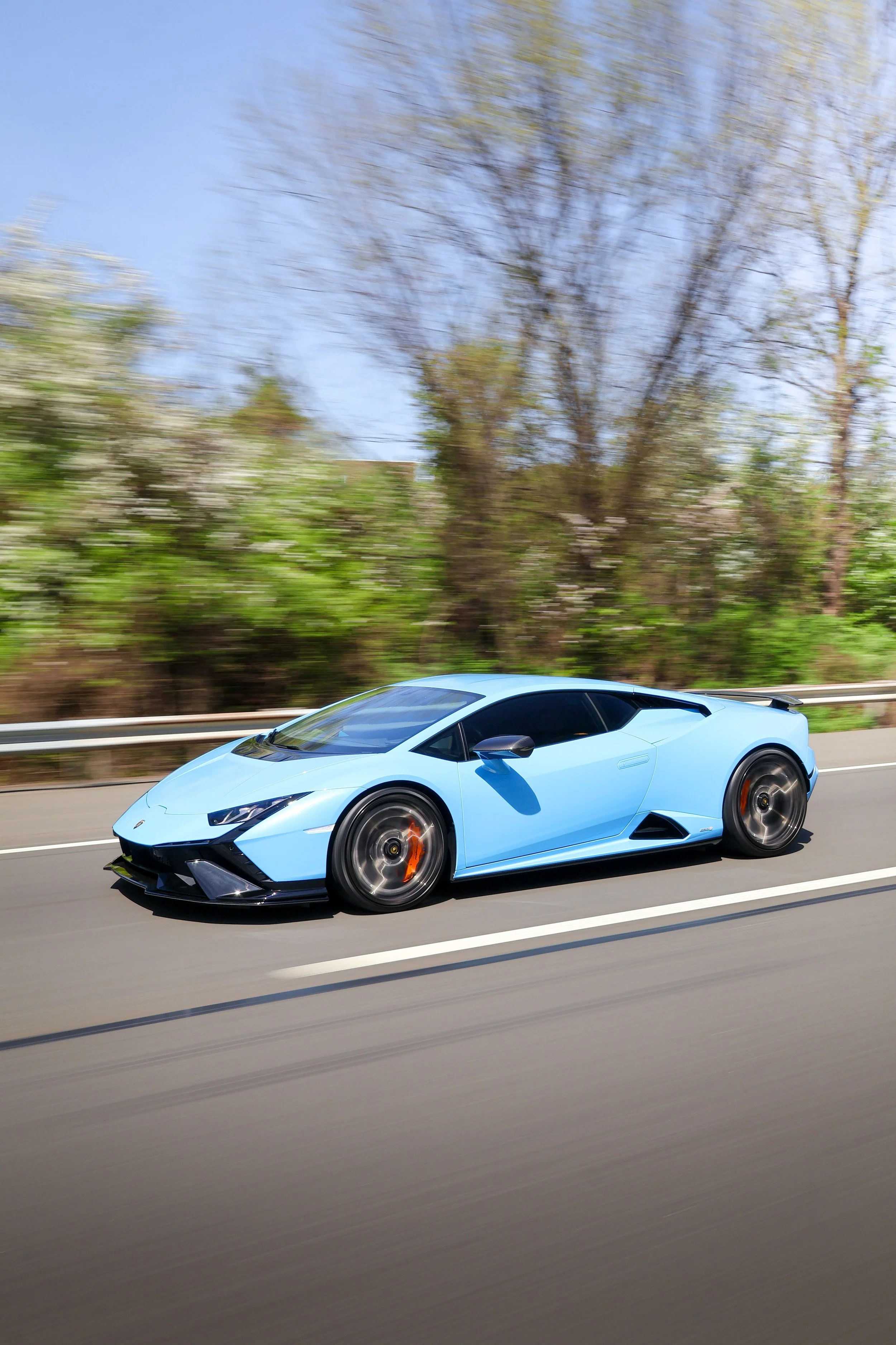 A light blue Lamborghini sports car is driving on a highway with trees and a blue sky in the background.