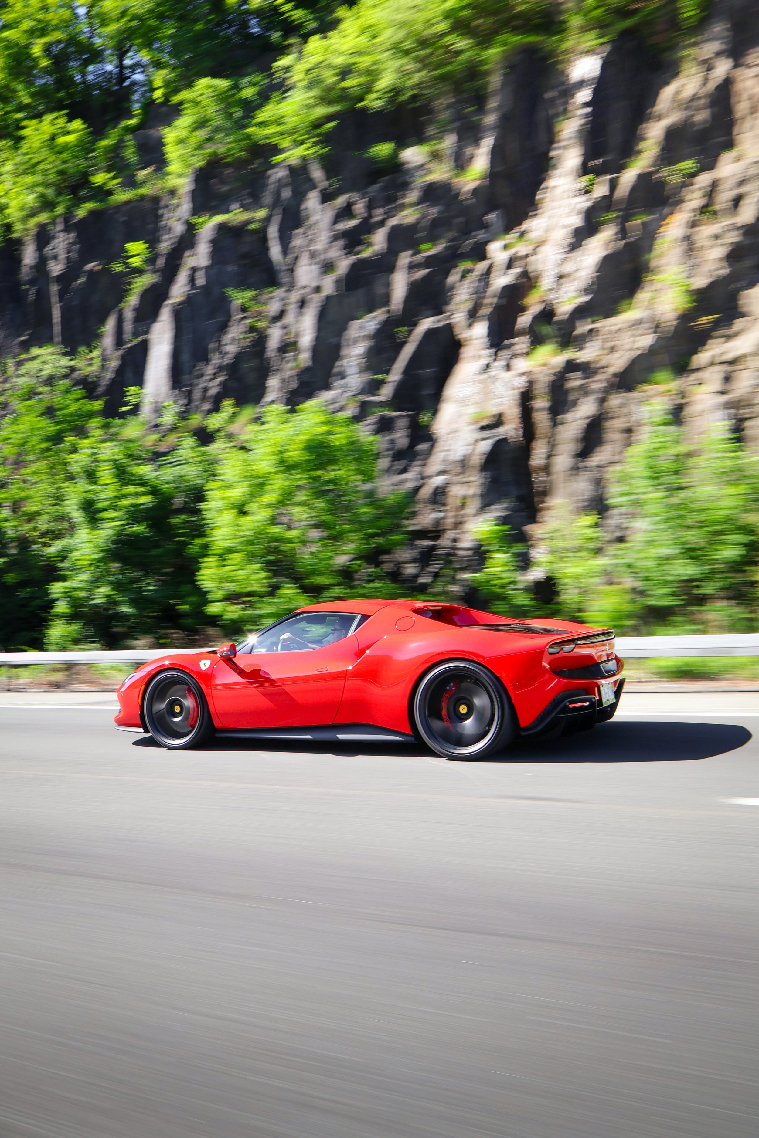 A red sports car driving on a highway with a rocky cliff and green trees in the background.