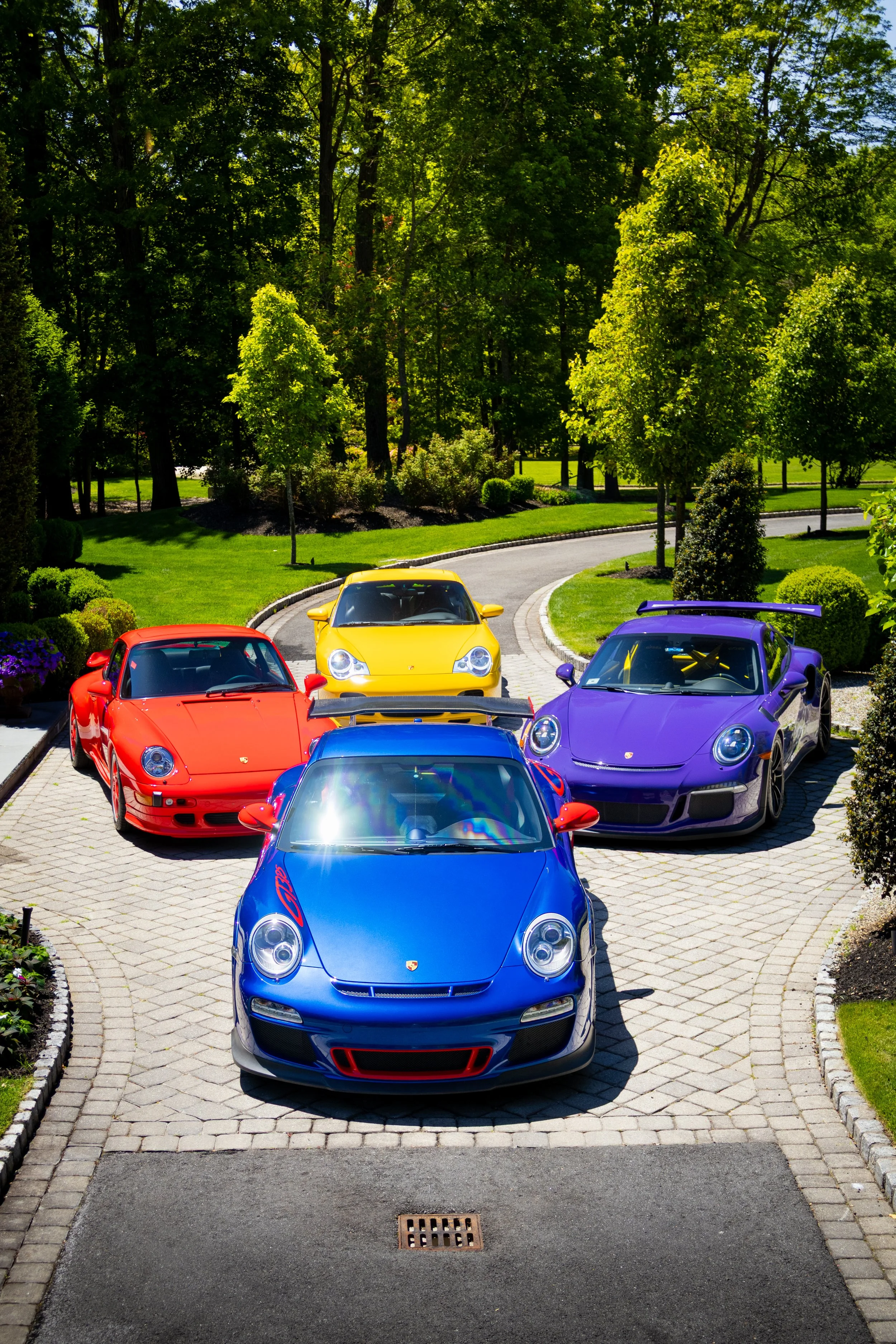 A row of colorful luxury sports cars, including a blue, purple, yellow, red, and another blue car, parked on a cobblestone driveway, with green trees and a well-maintained lawn in the background.