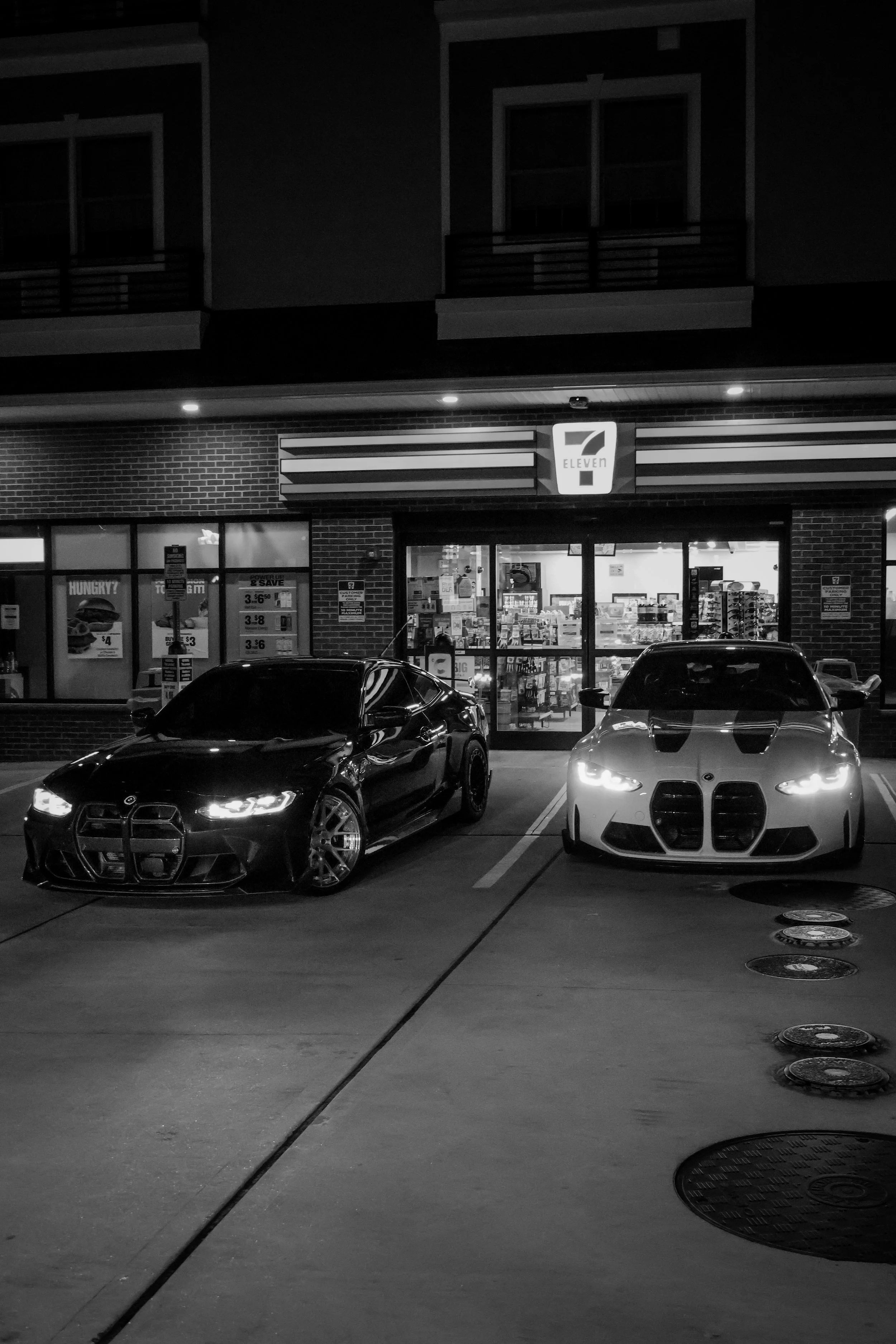 Nighttime scene of a parking lot with two cars parked in front of a 7-Eleven convenience store. The store's brightly lit sign is visible above the entrance, and inside the store, various products can be seen through the large glass windows.