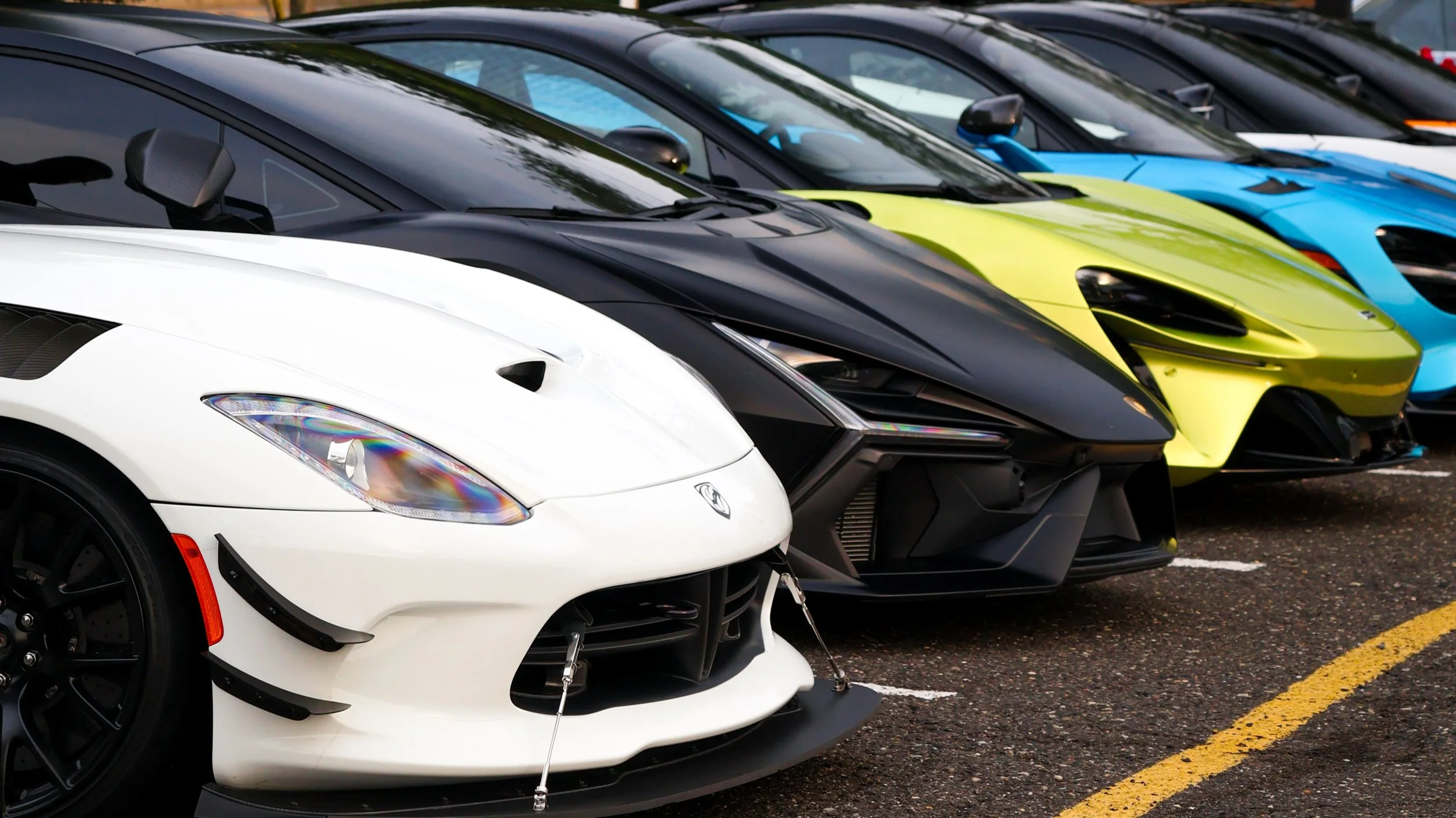 Row of luxury sports cars parked in a parking lot, including a white Dodge Viper, a black Lamborghini, a yellow Lamborghini, and a blue sports car, all with sleek designs.