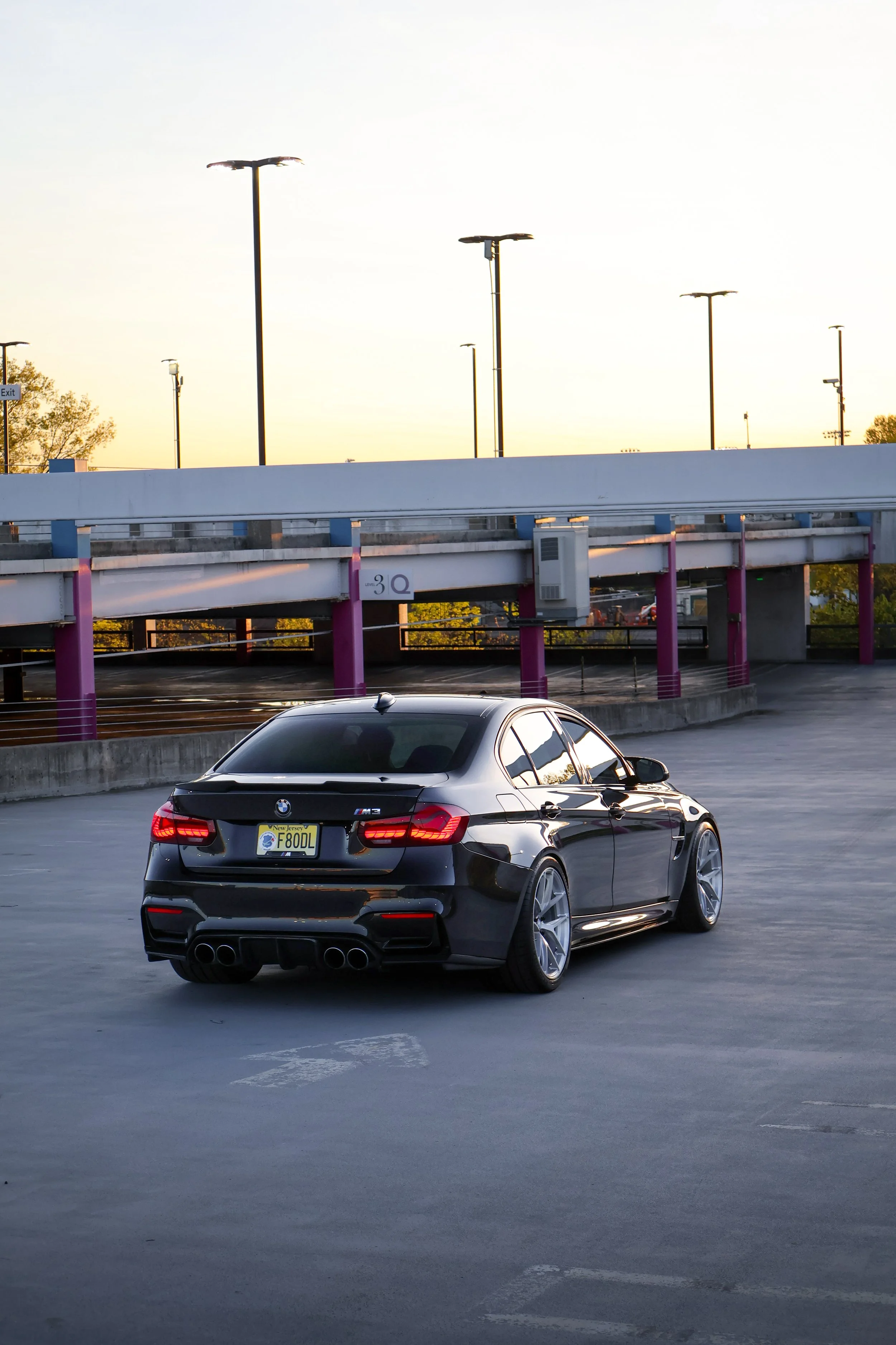 A black BMW M3 parked in an outdoor parking lot during sunset, with artistic pink and purple lighting accents on the parking structure.