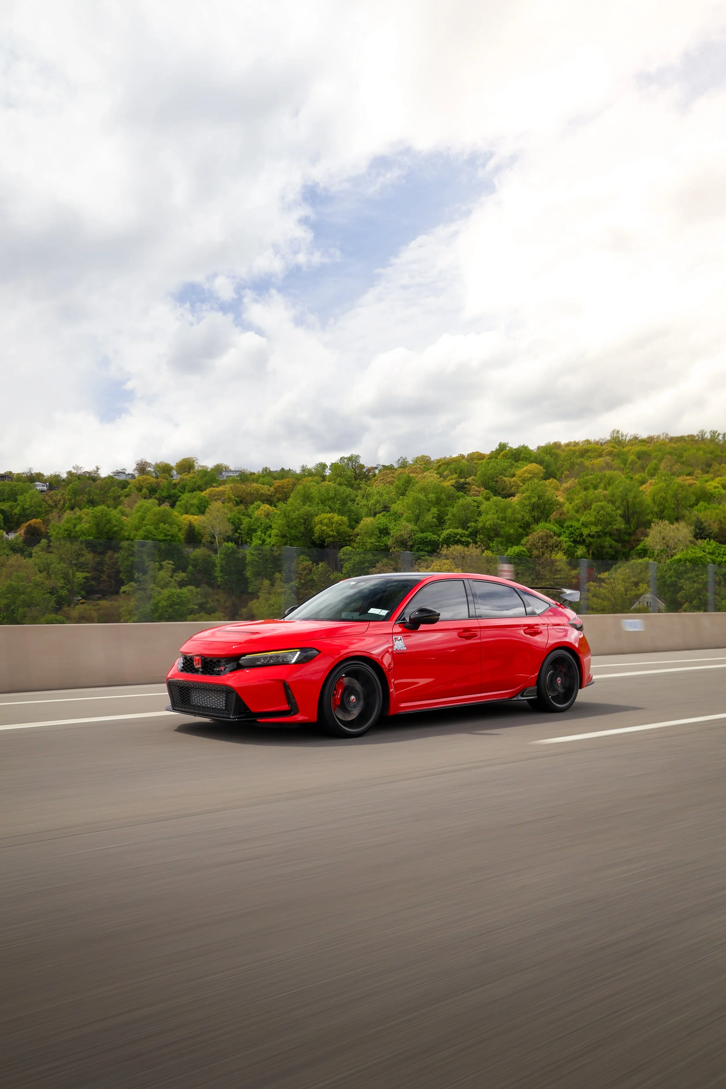 Red sports car driving on highway with green trees and cloudy sky in the background.
