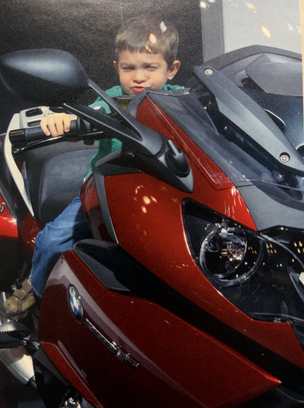 Young boy sitting on a red and black BMW motorcycle, making a face with puckered lips and furrowed brows, with one hand on the handlebar, inside a showroom or garage.