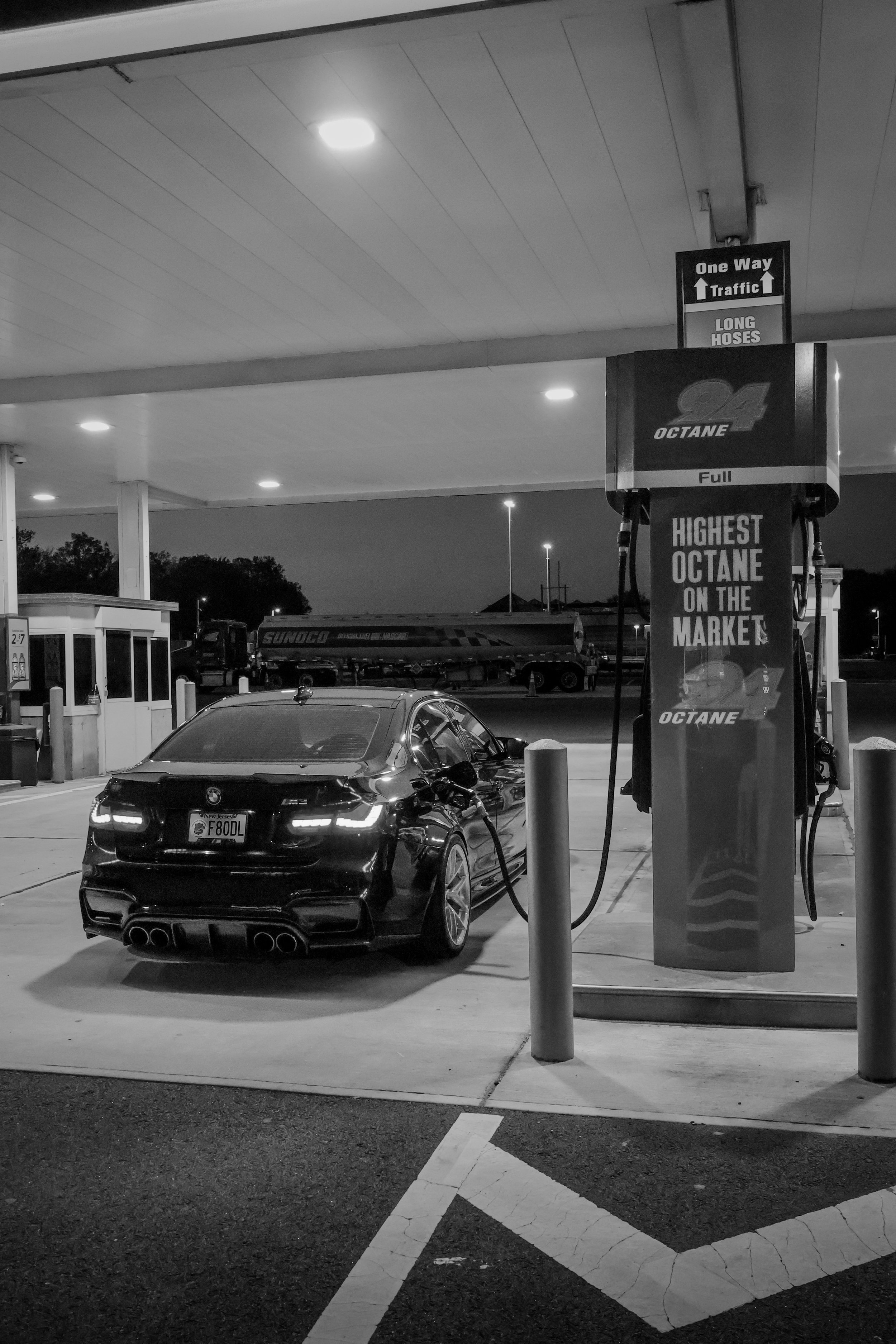 A black sports car parked at a gas station pump at night. The gas station has a canopy with ceiling lights, and in the background, there are more lights and trees. The pump displays 