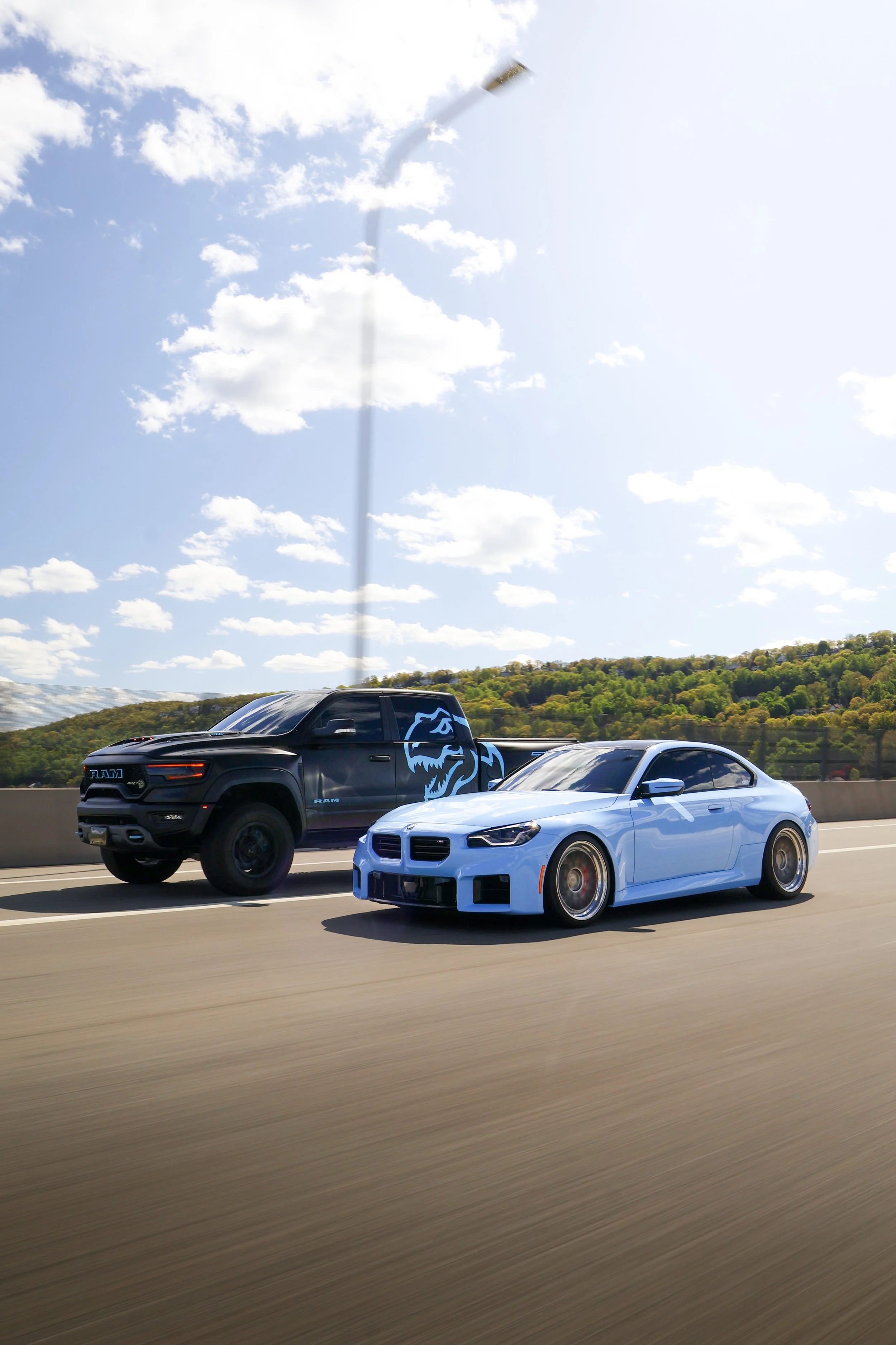 A black RAM truck with a blue lightning logo on the side and a white sports car driving on highway with green trees and blue sky with clouds in background.