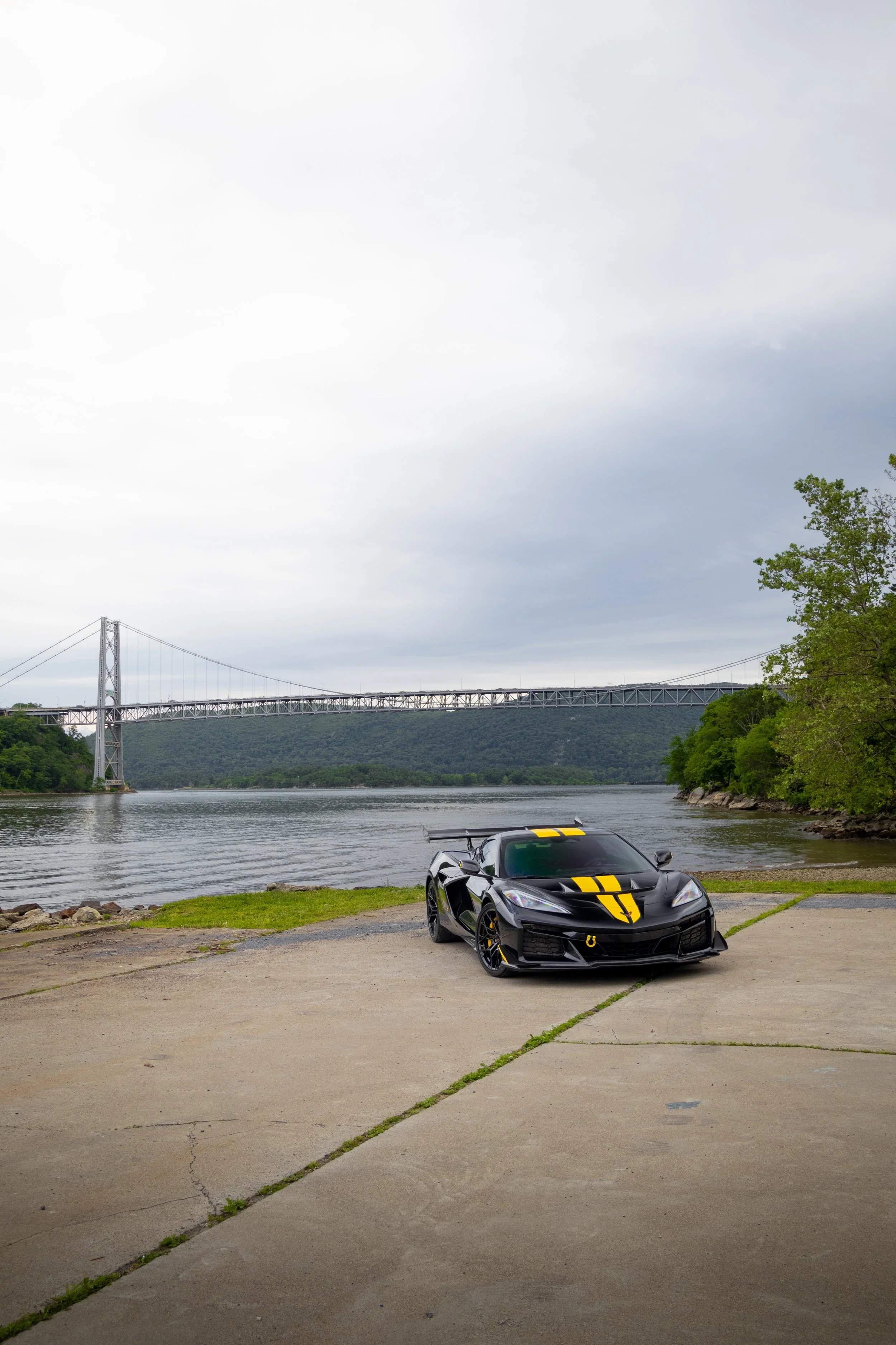 A sleek black sports car with yellow racing stripes parked on a concrete pathway near a river, with a large suspension bridge and green trees in the background under an overcast sky.