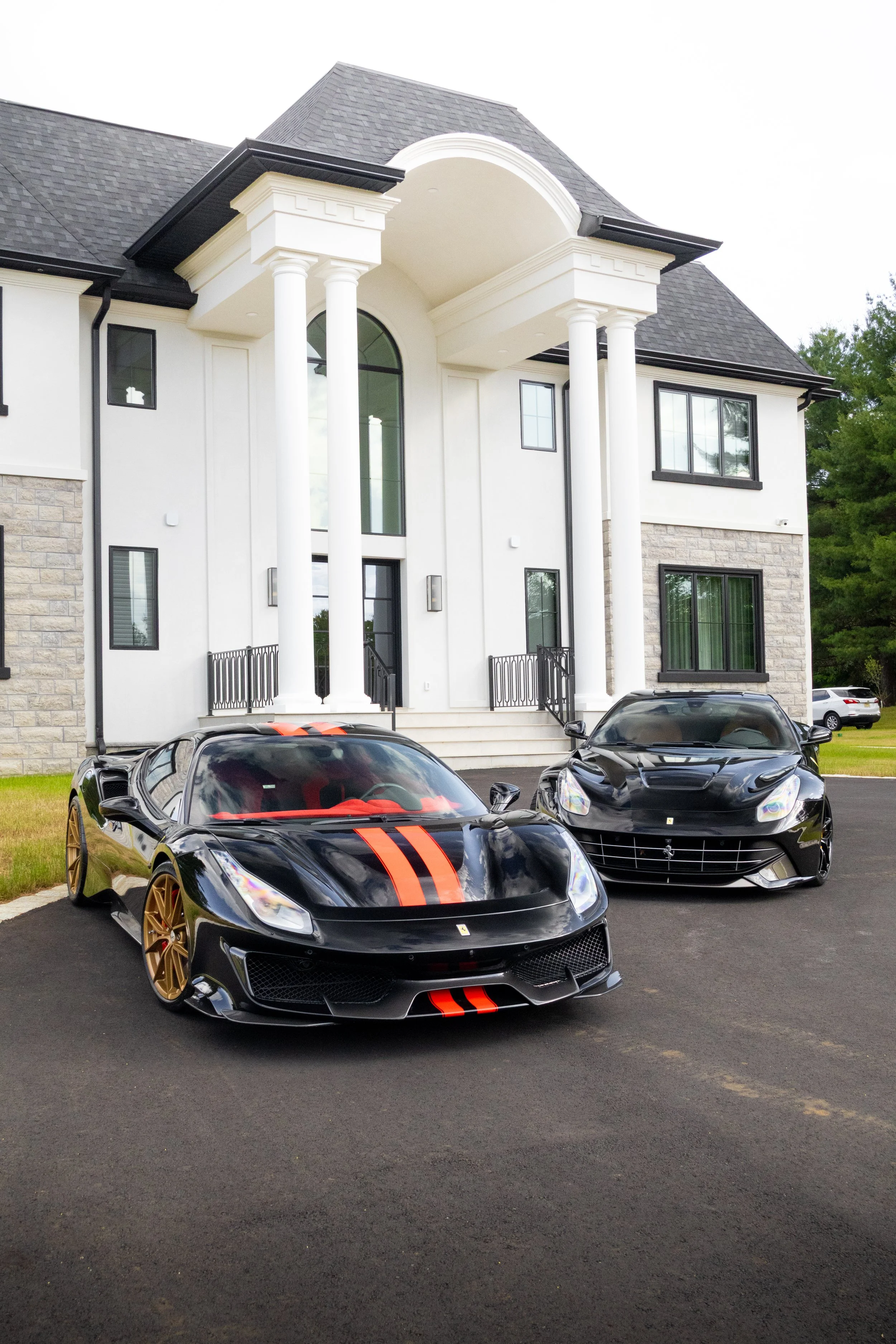 A black sports car with gold wheels and red racing stripes parked in front of a white, two-story mansion with large windows and decorative columns.