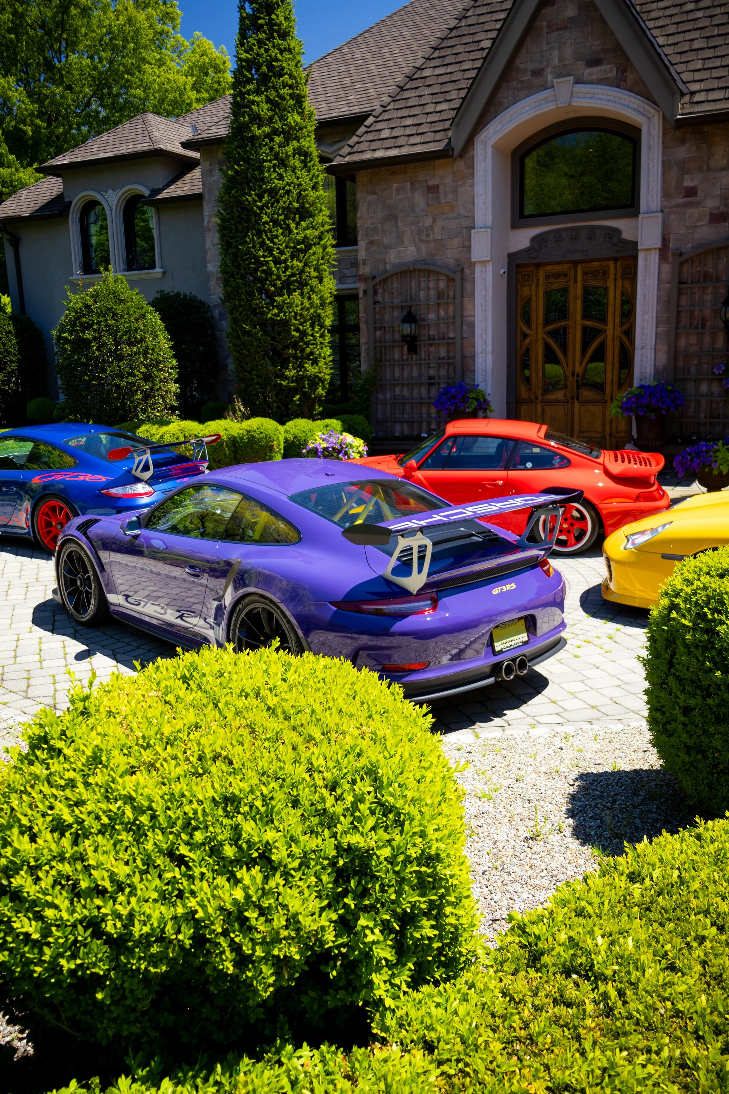 Colorful sports cars parked in front of a large house with lush green trees and neatly trimmed bushes.