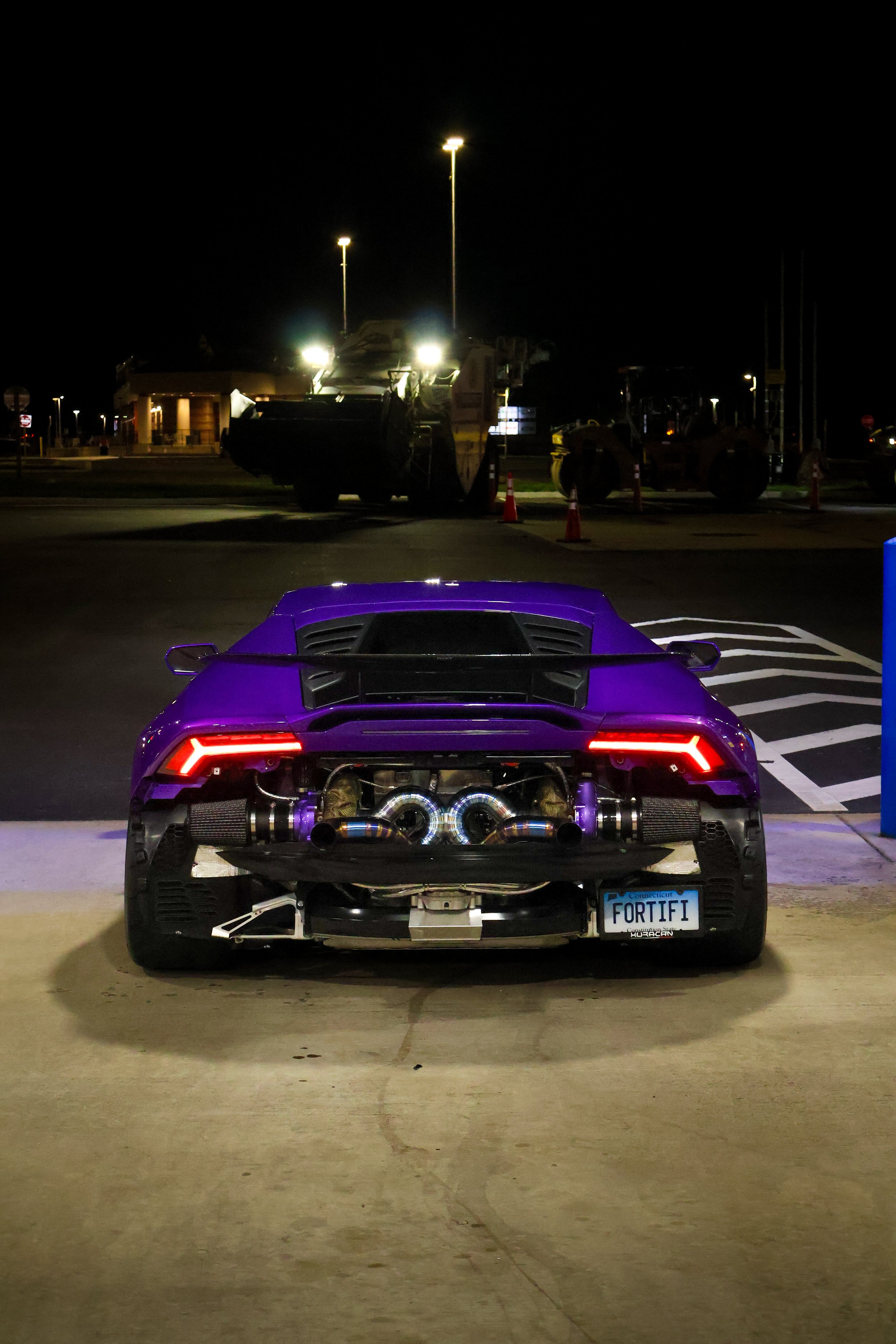 A purple sports car with visible exhaust pipes and a Connecticut license plate reading 'FORTIFI' parked in a lot at night. The background features construction equipment and streetlights.