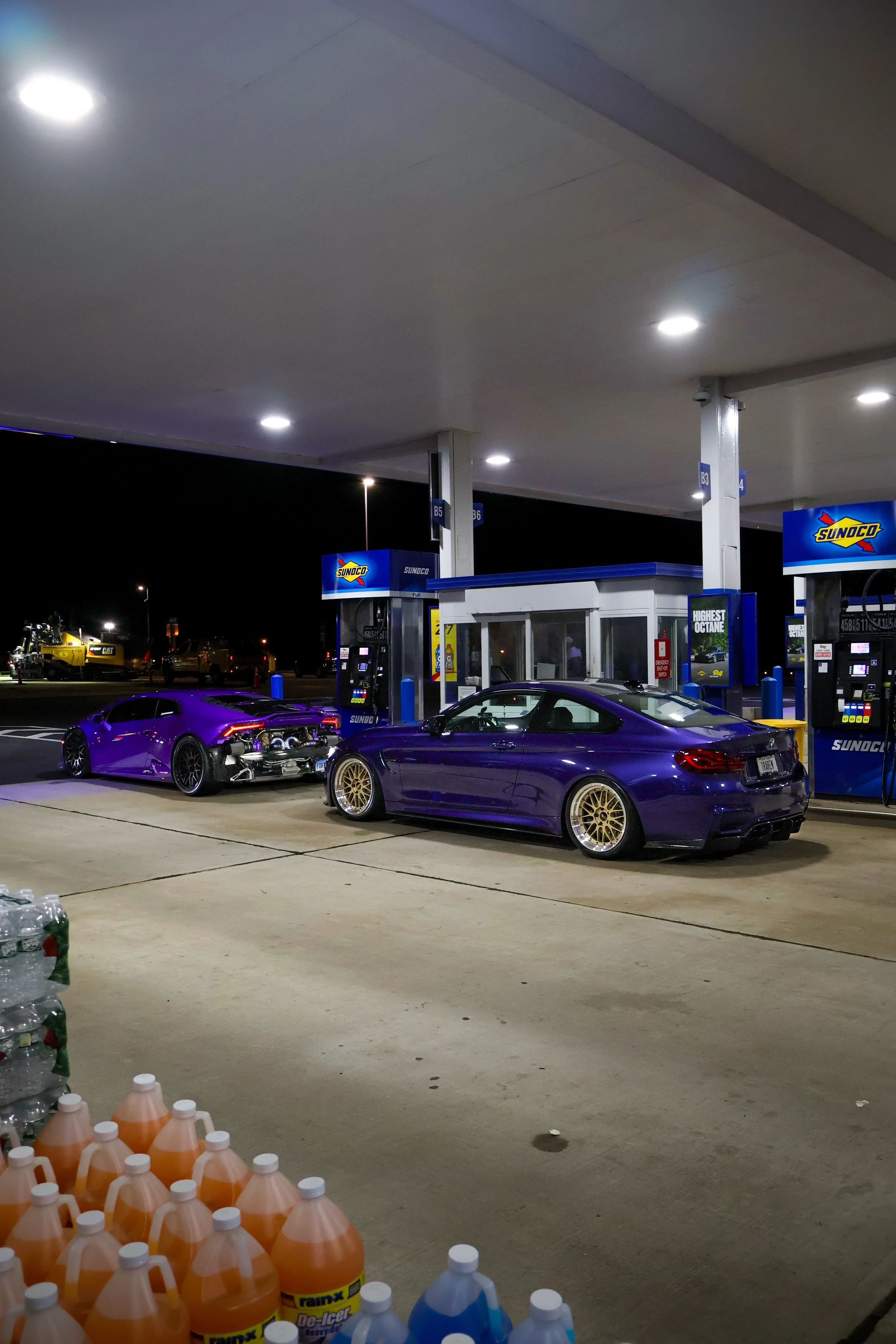 Nighttime scene at a Sunoco gas station with two luxury cars parked near the pumps. One car is purple and heavily modified with aftermarket wheels, the other is a dark purple or blue with gold wheels. There are fuel pumps and a small convenience stor
