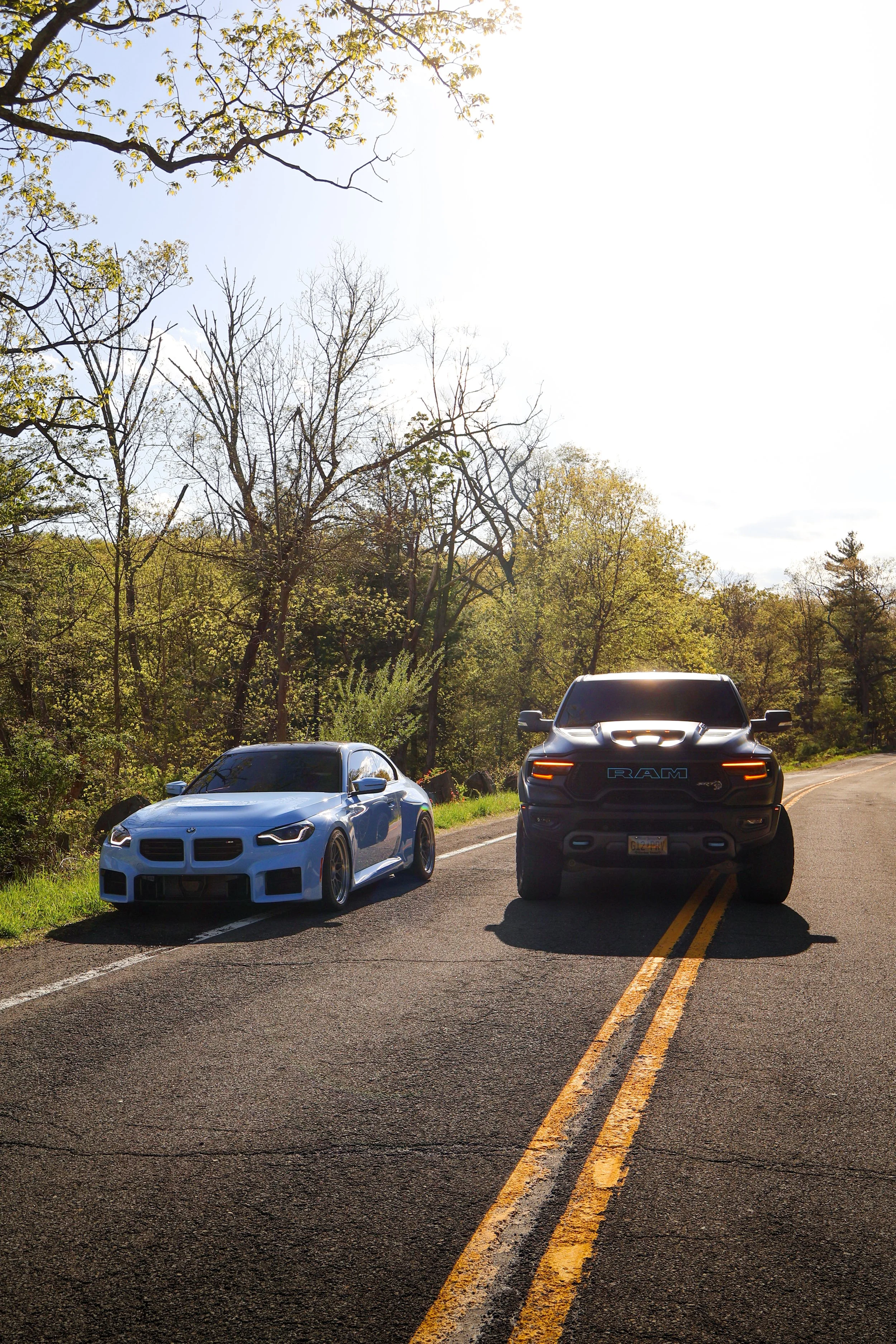 A blue sports car parked on the left and a black pickup truck parked on the right on a rural road with double yellow lines. There are trees with emerging leaves and partly cloudy sky in the background.