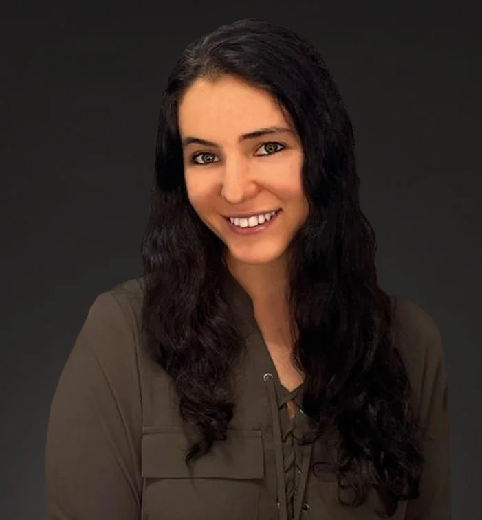A woman with long, dark wavy hair smiling at the camera against a dark background.