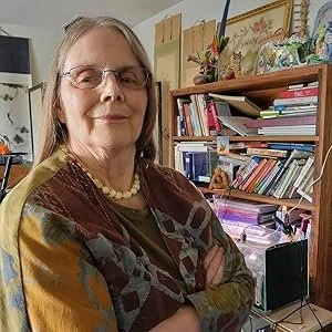 An older woman with glasses and shoulder-length hair standing in a home office surrounded by books and decorative items on shelves.