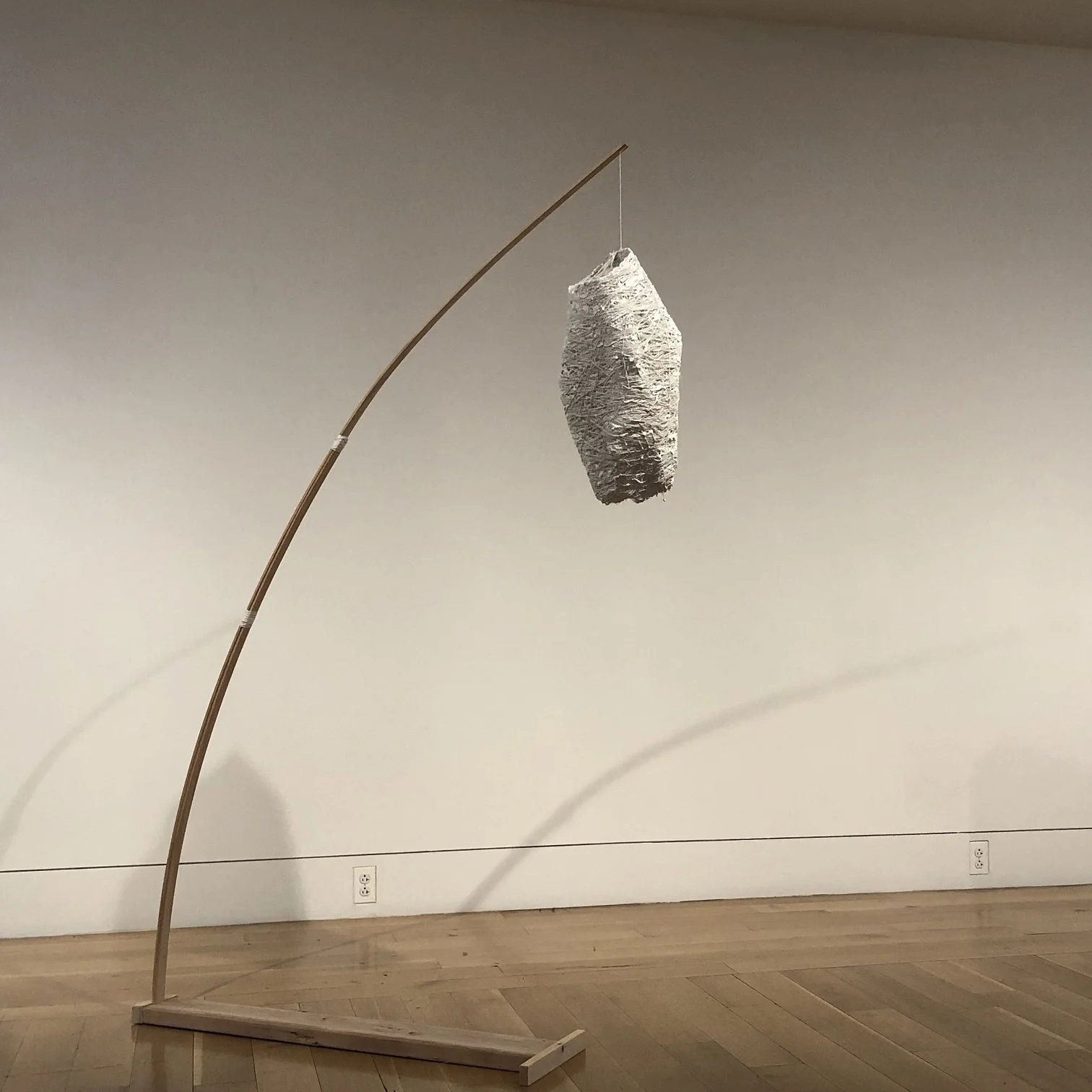 A modern art sculpture featuring a bent wooden stand supporting a textured paper or fabric bundle, hanging from a thin string, against a plain wall.