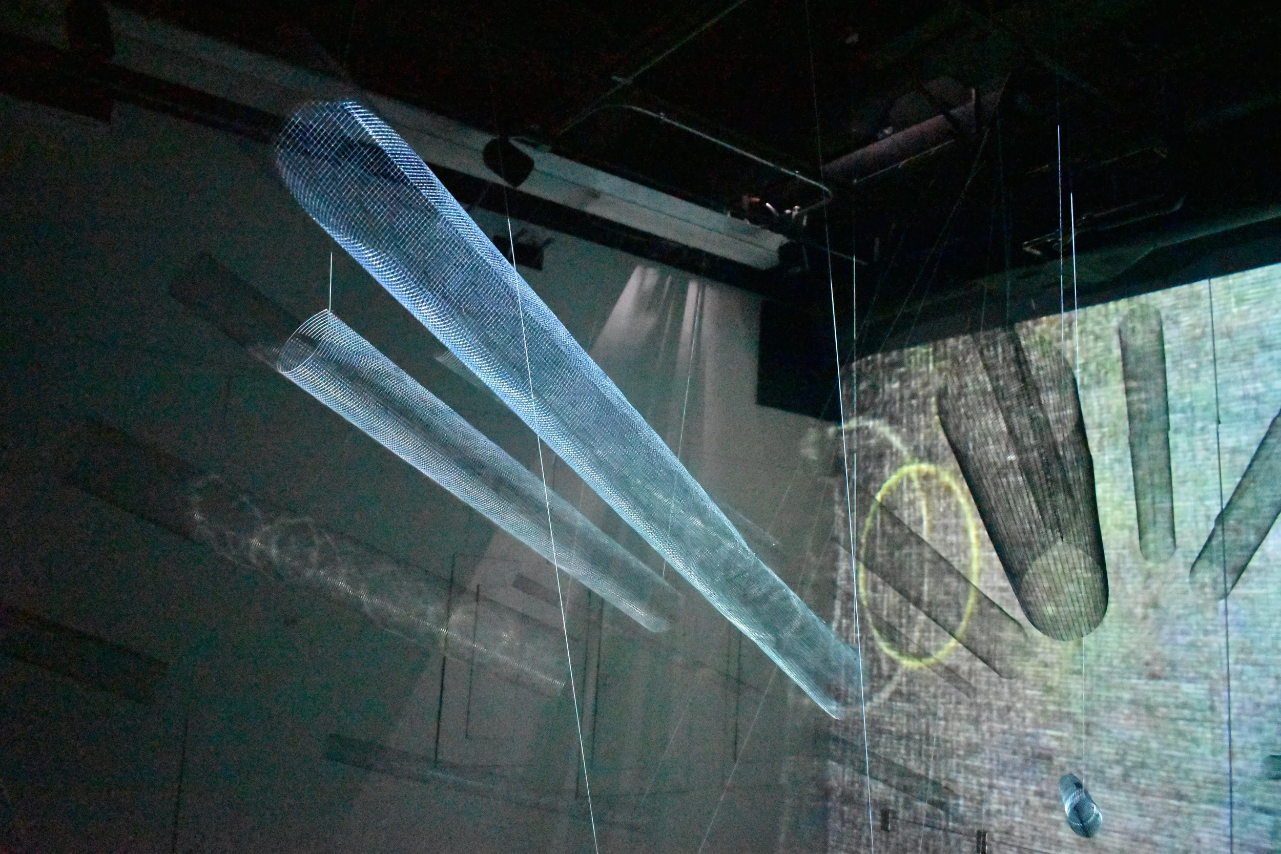 Installation art featuring translucent cylindrical structures with a grid pattern, suspended from the ceiling with wires in a dark gallery space, with a large screen displaying an outdoor scene in the background.