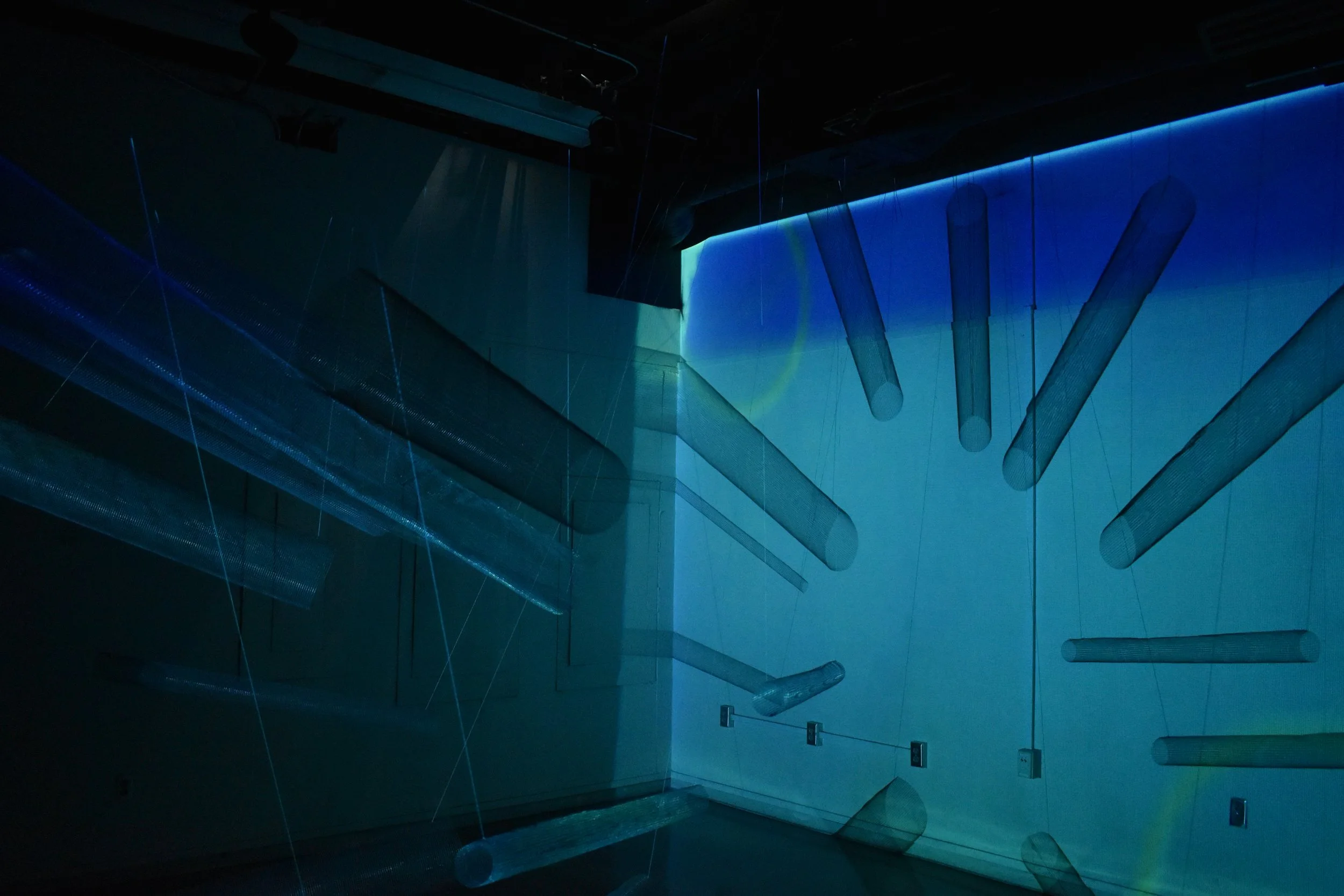 An abstract art installation with hanging cylindrical and rectangular mesh-like structures illuminated in blue and green light, creating a surreal, space-like atmosphere.
