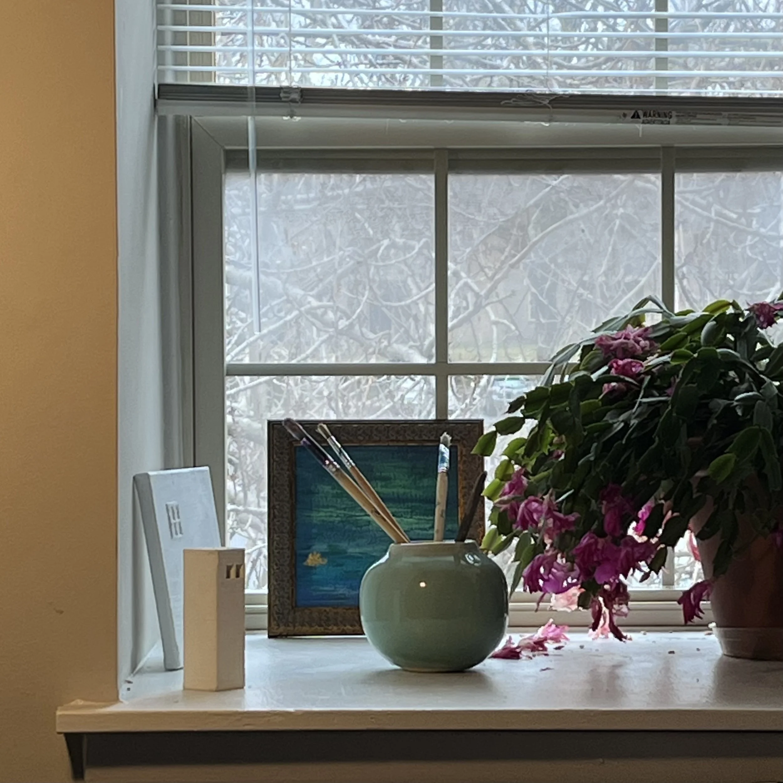 A windowsill with pink flowering plant, a green vase with paintbrushes, a framed painting of a landscape, and two decorative books, with a snowy scene outside.