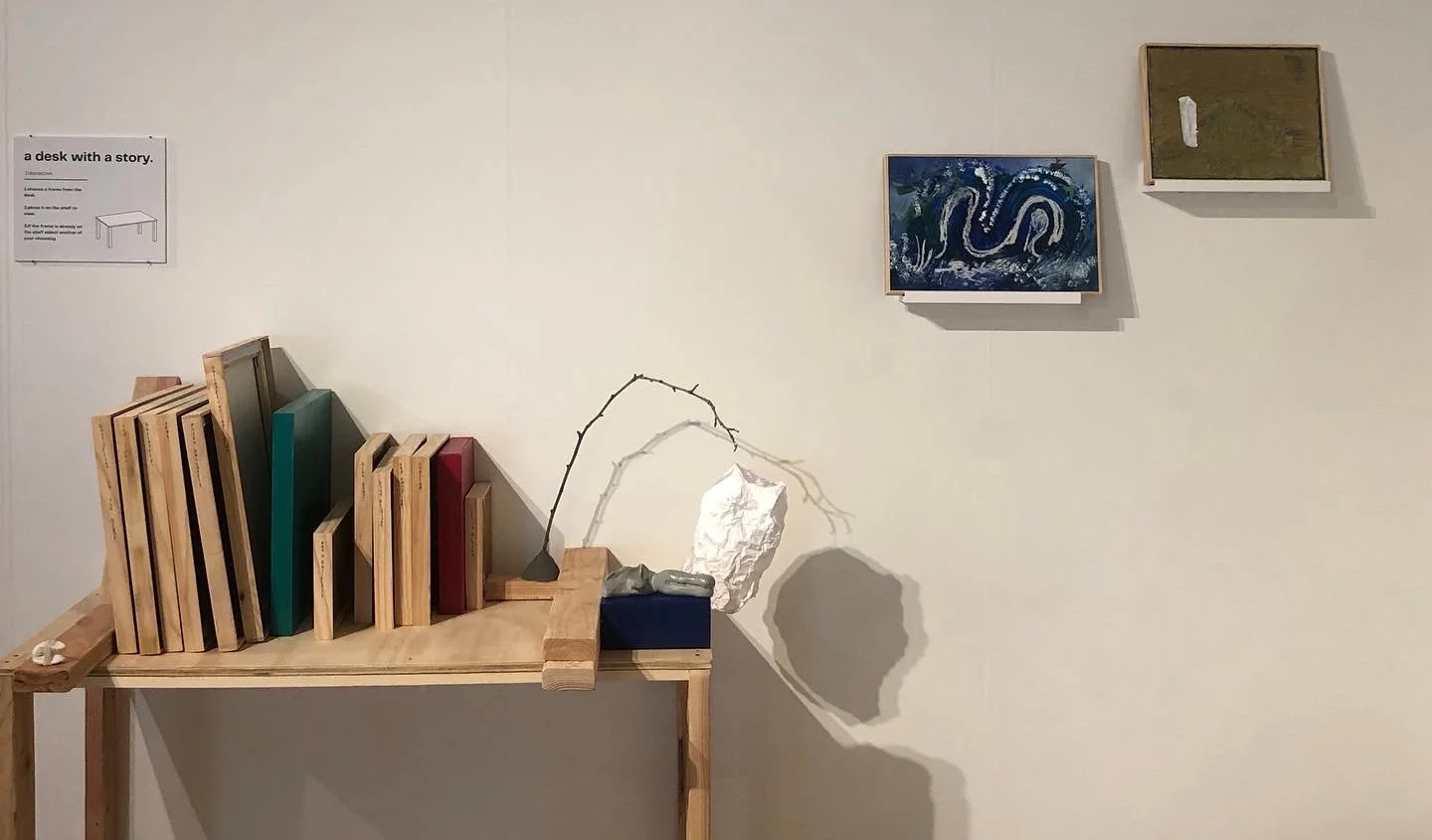 Art installation featuring books, a branch, and a paper sculpture on a wooden table, with framed artworks on a white gallery wall.